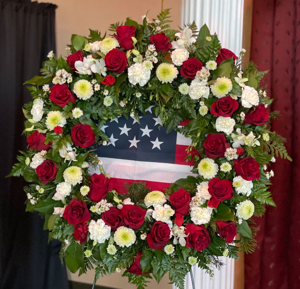 Wreath WR-02 Wreath WR-02 - This patriotic wreath pays tribute with a bold ring of red roses, white carnations, green-centered mums, and accent blooms, all set against a lush bed of greenery. The American flag is carefully placed at the center, offering a proud and heartfelt farewell. Easel stand included.