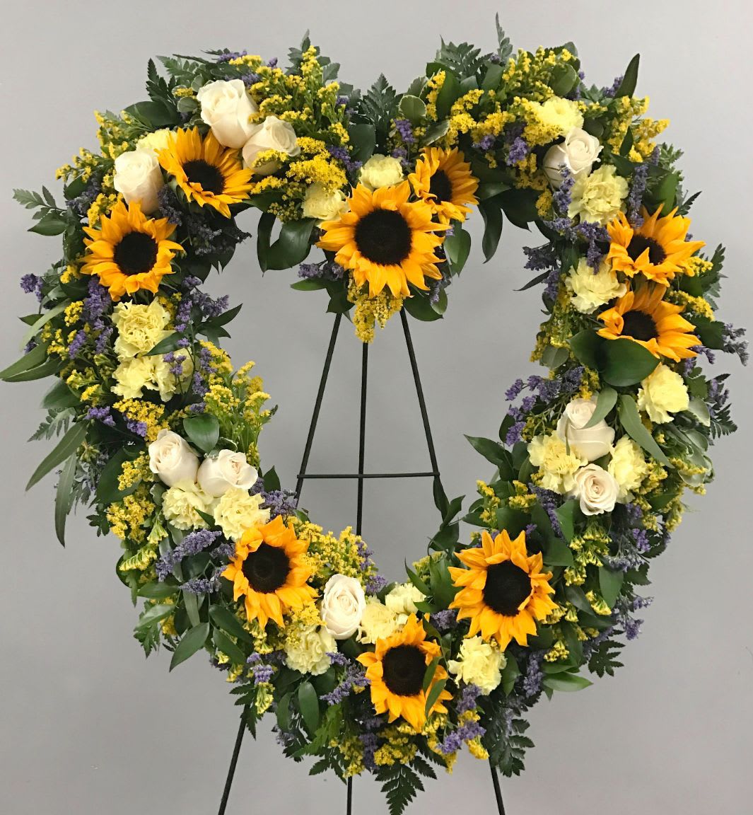 Heart HS-01 Heart HS-01 - This heart-shaped standing wreath offers a warm and uplifting tribute with golden sunflowers, soft yellow carnations, ivory roses, and accents of lavender misty and solidago. Its cheerful yet heartfelt design is a beautiful way to celebrate a vibrant life, especially for services in Litchfield Park, Buckeye, and Goodyear. Easel stand included; ribbon lettering available for $10 per word.