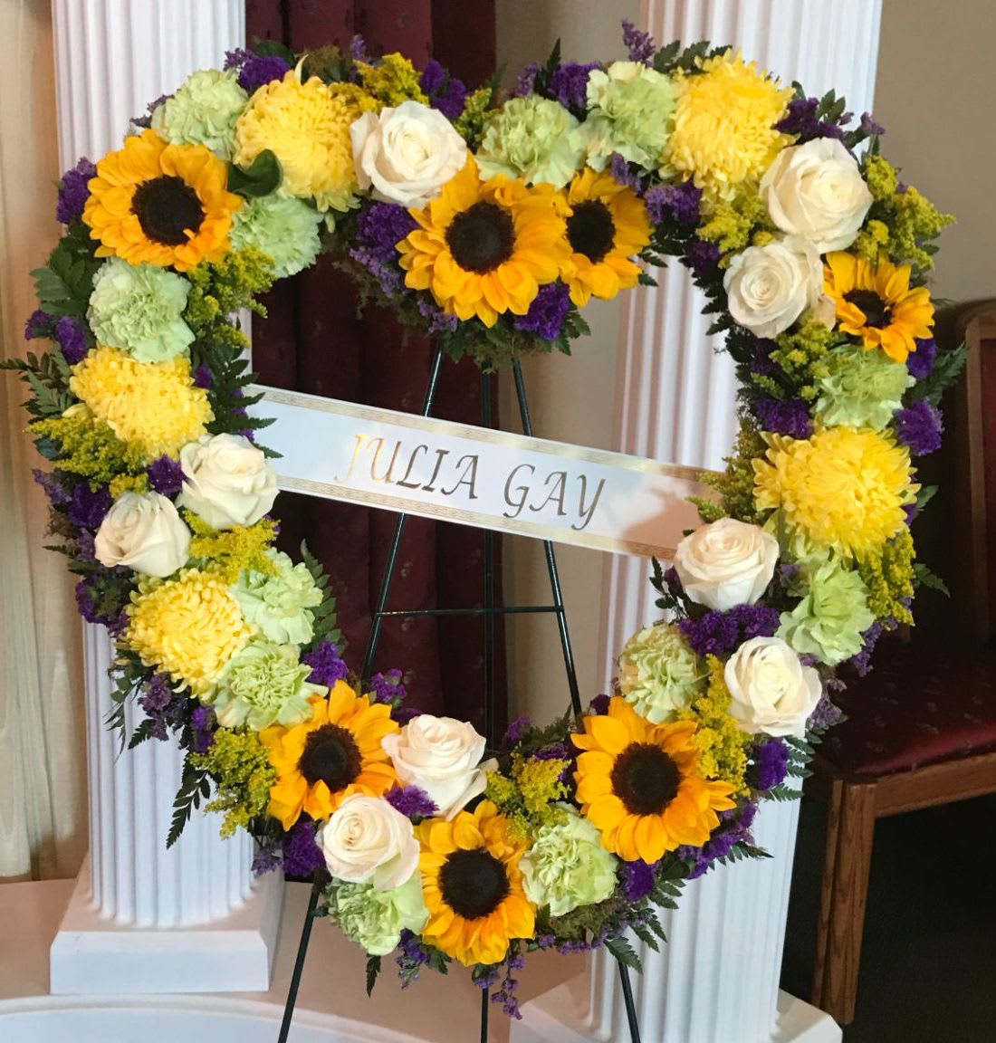 Heart HS-09 Heart HS-09 - This heart-shaped wreath is filled with cheerful sunflowers, soft white roses, bright yellow mums, and fresh green carnations, accented with touches of purple statice and solidago. Its warm, garden-inspired color palette brings comfort and light to the service. A fitting tribute for memorials held in Estrella Mountain Ranch, Goodyear, or Avondale. Easel stand is included; ribbon can be added for $10 per word.