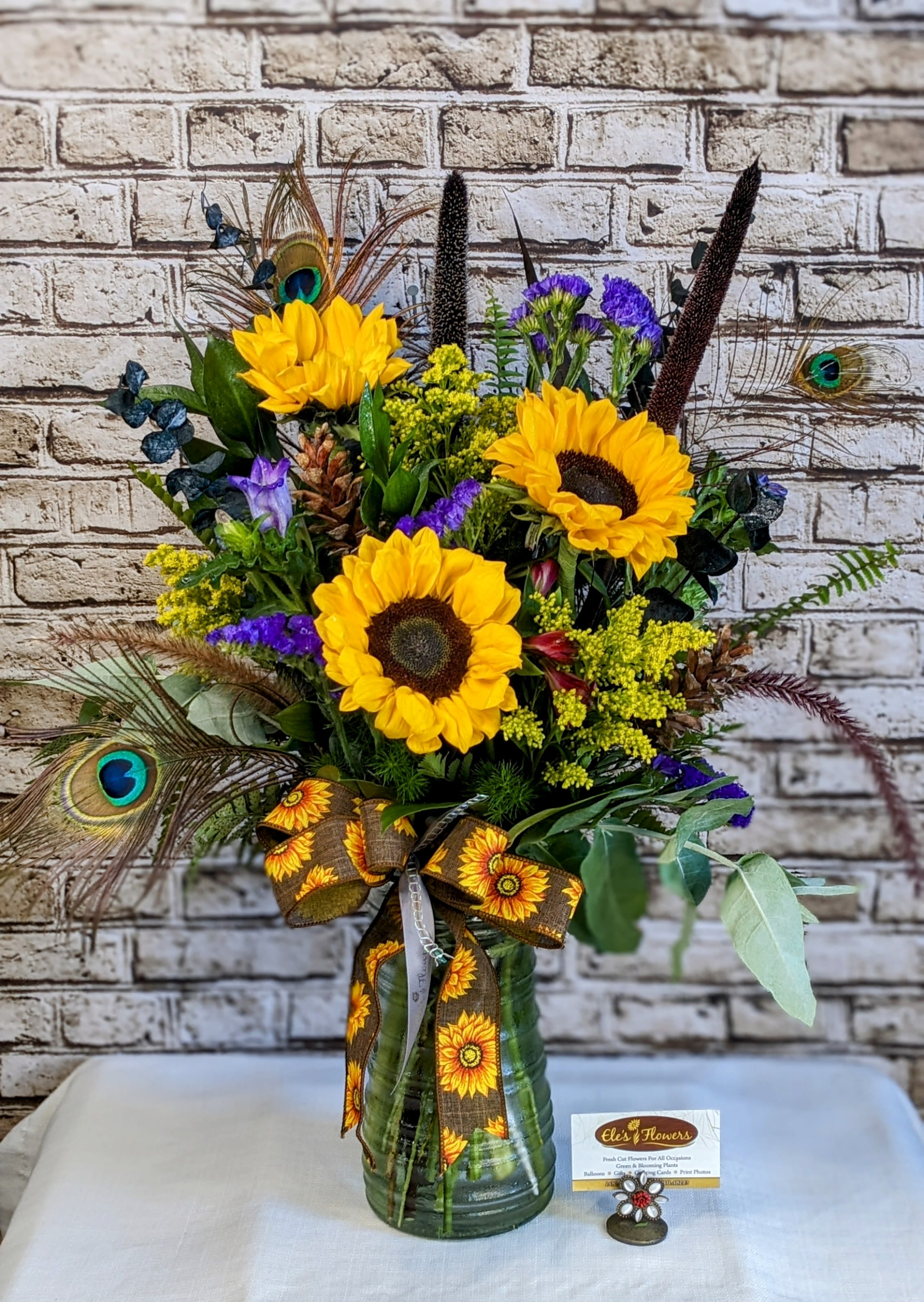 Golden Radiance Golden Radiance - Bring a burst of sunshine indoors with this bold and cheerful sunflower arrangement. Featuring radiant yellow sunflowers paired with rich purple blooms, golden solidago, eucalyptus, and playful textures, this design is both vibrant and uplifting. Unique accents like peacock feathers, pinecones, and tall millet stems add a touch of rustic charm and artistic flair. Arranged in a clear glass vase, the bouquet is finished with a sunflower-themed ribbon, making it a perfect gift for any occasion—whether to brighten someone’s day, celebrate a special milestone, or add a pop of seasonal color to your home. Approximate size: 28–30"H x 20–22"W
