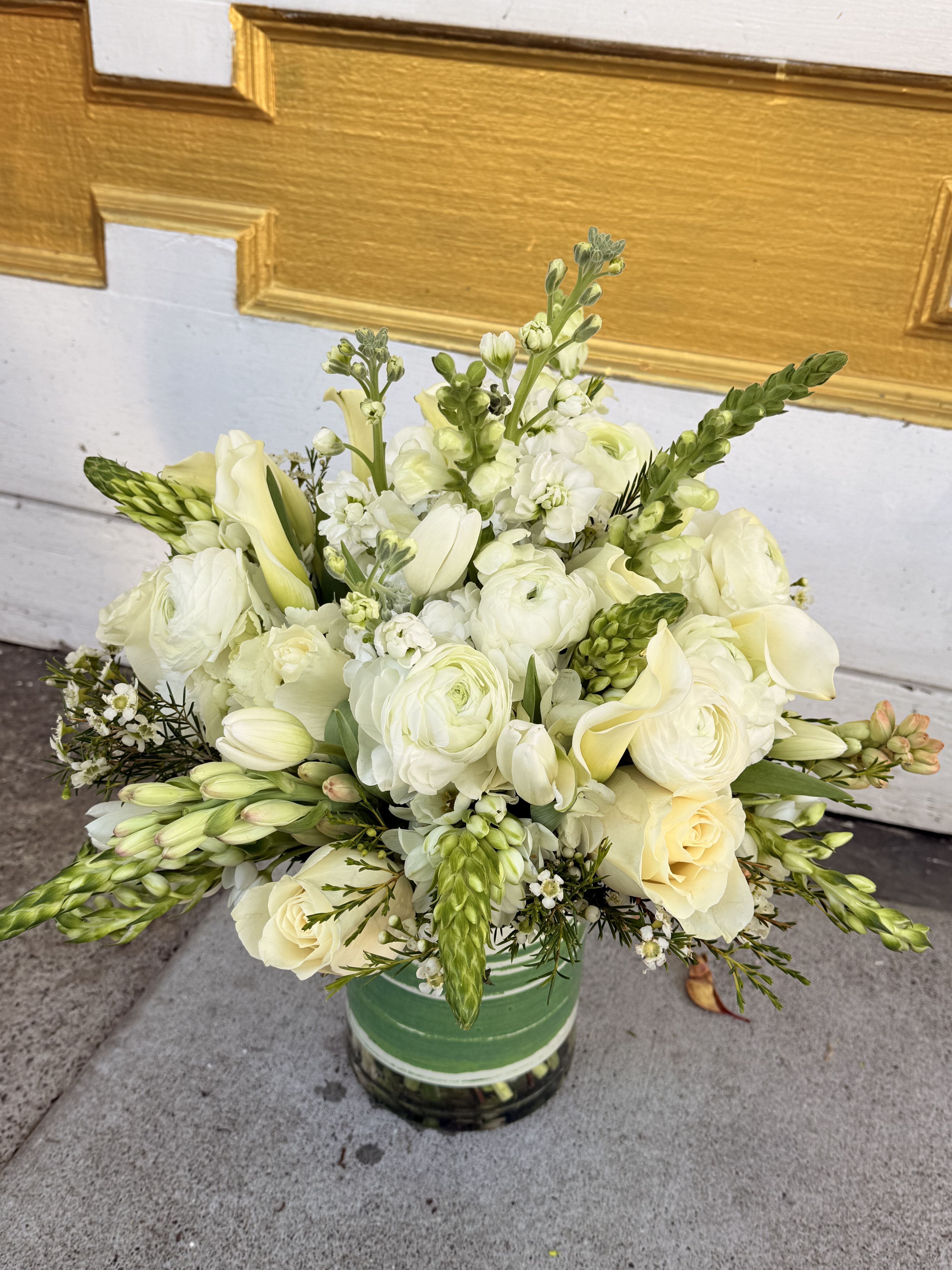 Ivory Whisper - This sophisticated arrangement features a harmonious blend of creamy whites, soft ivories, and fresh greenery, creating an elegant and timeless look. The bouquet includes: 	•	White ranunculus – adding lush, layered texture 	•	Cream roses – soft and romantic focal blooms 	•	Calla lilies – graceful and modern accents 	•	Tuberose buds – introducing delicate fragrance and vertical movement 	•	White stock or lisianthus – adding volume and soft ruffles 	•	Wax flowers – tiny, dainty fillers enhancing texture 	•	Green buds and foliage – adding freshness and balance  Arranged in a clear glass vase wrapped with a green leaf lining, this design is perfect for sophisticated occasions — weddings, anniversaries, or an elegant sympathy gesture. Its neutral palette makes it versatile and timeless.