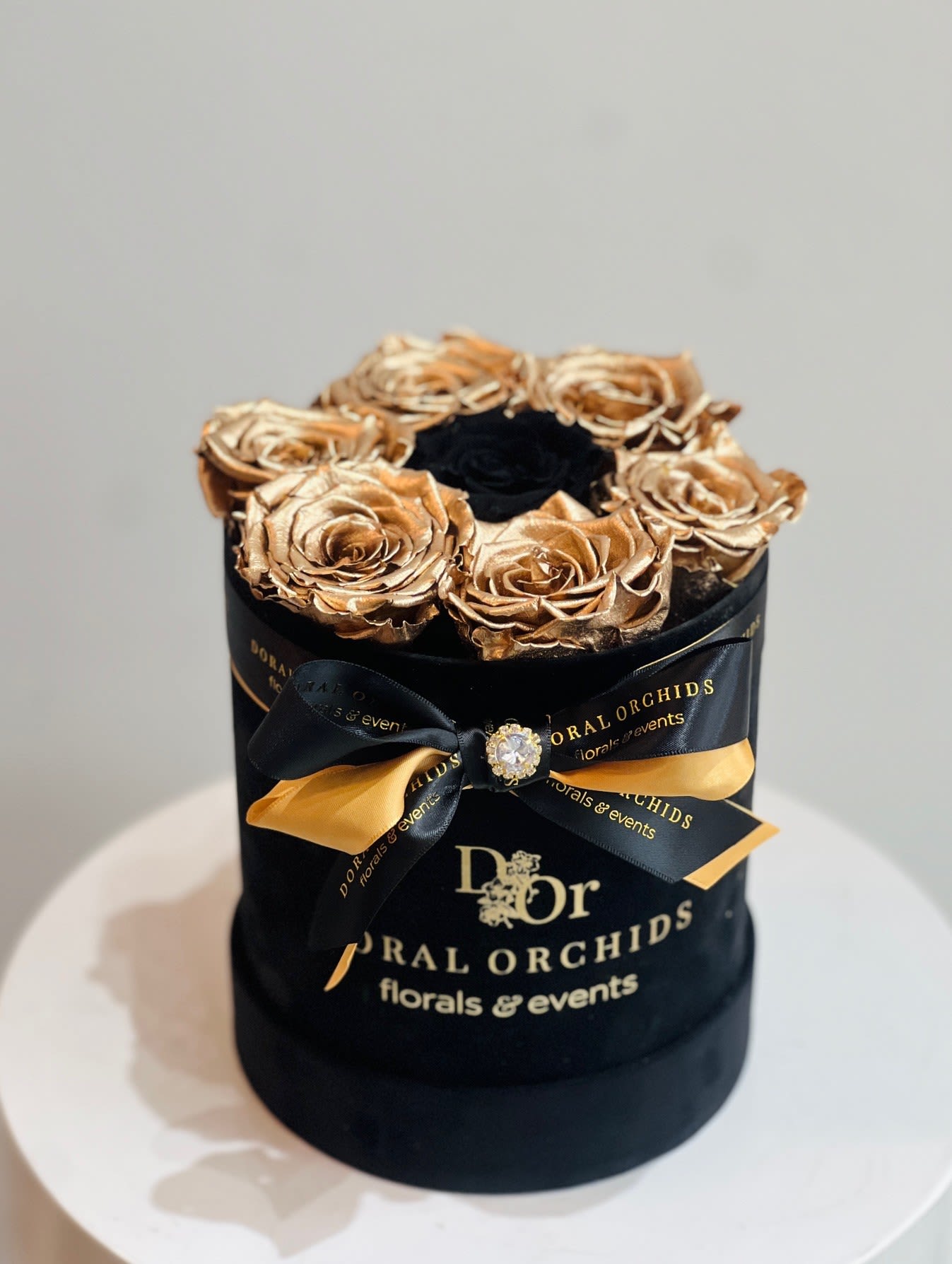 Lovely Roses - Black and Gold - This luxurious arrangement showcases natural preserved roses—metallic gold blooms encircling a dramatic black rose at the center. Specially treated to last 3 or more years, they are presented in a black velvet hatbox with a satin bow and jewel accent, creating a bold and timeless display of elegance.