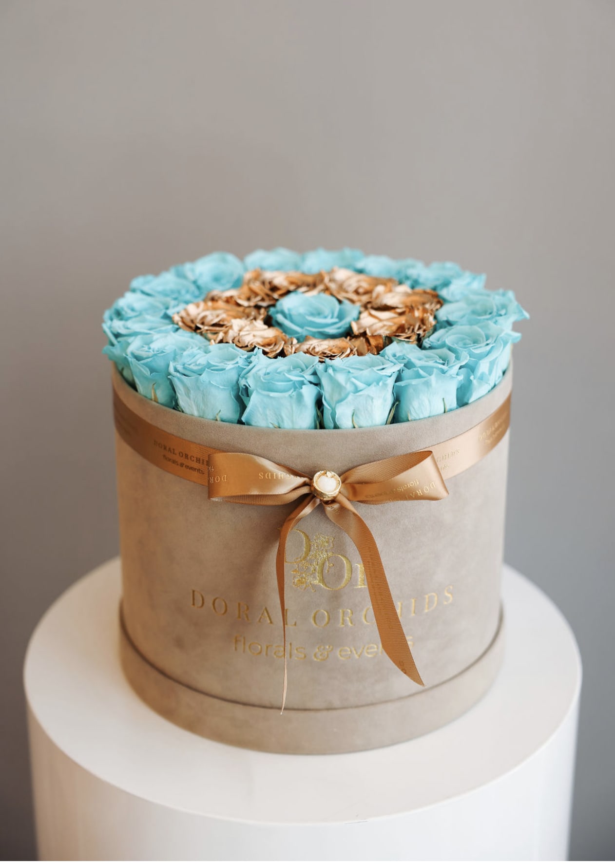 Lovely Roses - aqua &amp; gold ™ - An opulent ring of shimmering gold roses encircles preserved aqua blooms, crowned with a central rose. This 3 or more years lasting arrangement in a champagne velvet hatbox exudes grandeur and modern elegance.