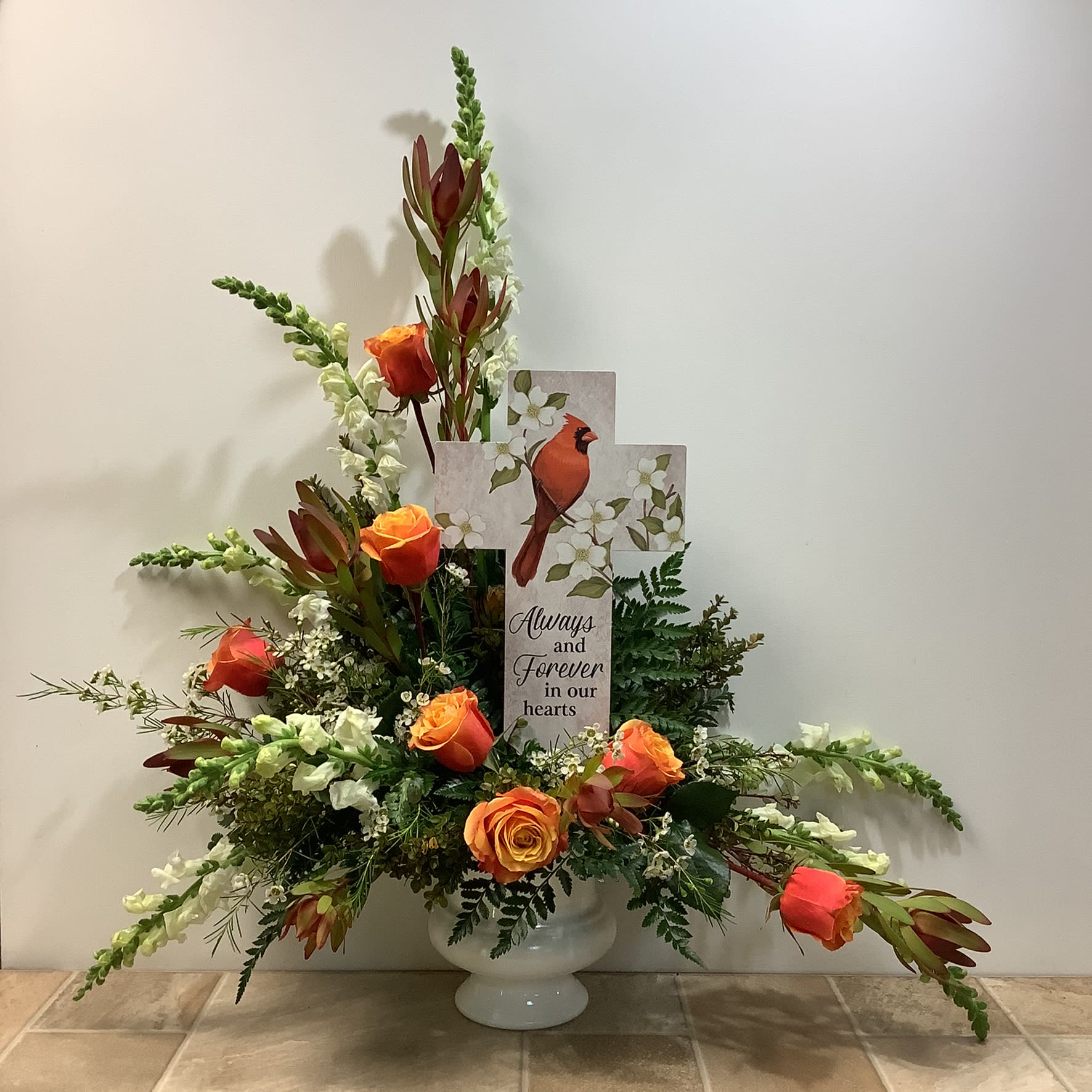 Always and Forever Bouquet Always and Forever Bouquet - "Always & Forever" is a graceful and heartfelt tribute arrangement designed to offer comfort and remembrance. It is artfully presented in a classic white pedestal, urn-style vase. At the center is a decorative cross-shaped garden stake plaque featuring a vibrant red cardinal perched among soft white blossoms. The cross bears the touching message: “Always and Forever in our hearts.” Cardinals, often seen as symbols of loved ones who have passed, add deep emotional resonance. The flowers include orange and sunset-toned Cherry Brandy roses which add warmth and symbolize admiration and love, creamy white snapdragons to represent strength and grace, rusty red leucadendron for an earthy texture, and charming white waxflowers. Lush greenery enhance the arrangement's fullness. It measures approximately 30” wide x 36” tall. Primary flowers: Rose, snapdragon, waxflower