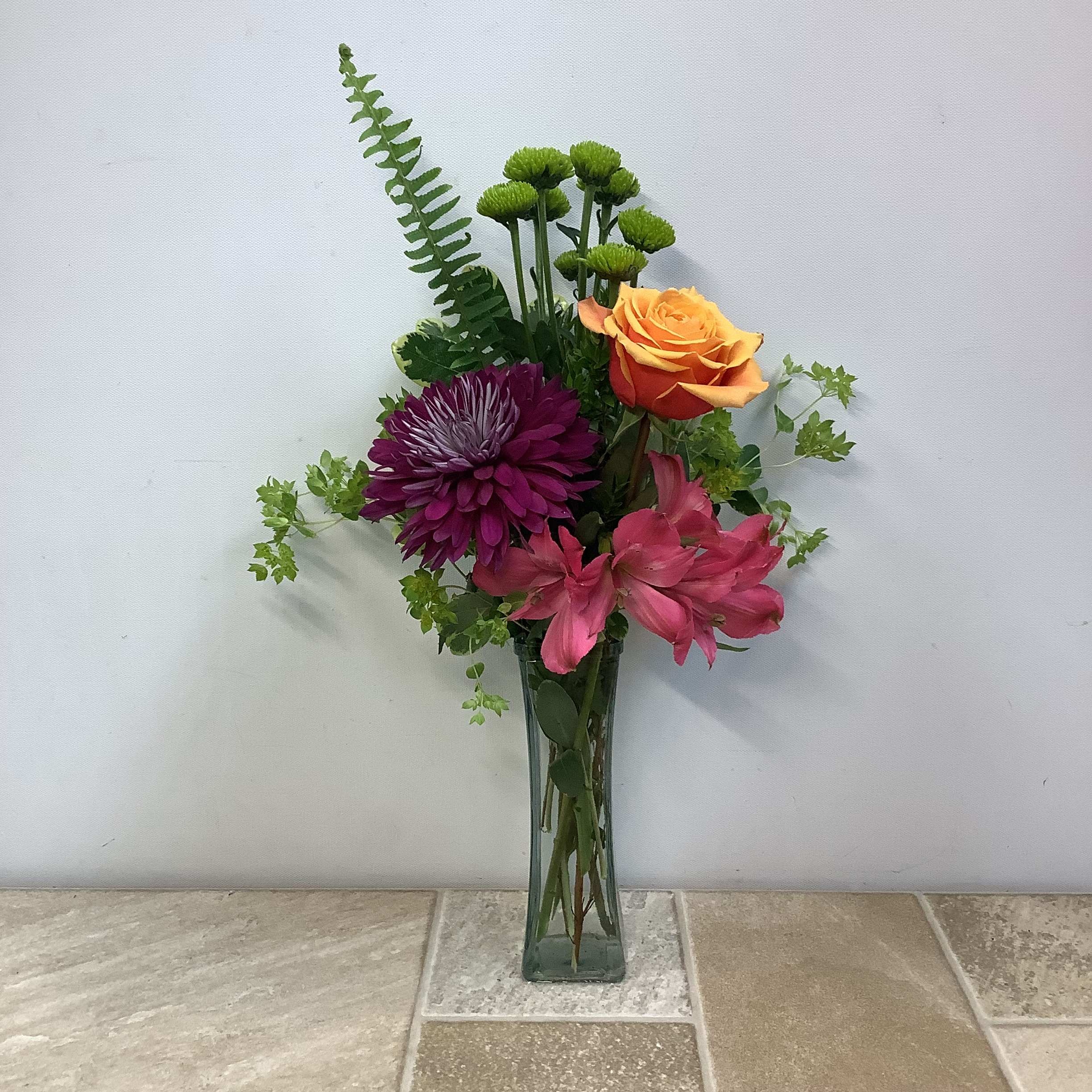 Annie’s Bouquet  - This bright and cheerful bud vase arrangement features a vibrant mix of colors and textures. &quot;Annie's Bouquet&quot; includes a gorgeous, orange-ish Cherry Brandy rose, adding a warm and cheerful focal point; a plum-colored chrysanthemum, providing bold contrast and rich color; pink alstroemeria, offering a touch of playfulness and movement; and bright green button poms, adding a fresh and whimsical accent. Assorted greenery lends  texture, height, and a natural feel to the design.  All flowers are arranged in a tall, slender, clear glass vase, creating a clean and contemporary presentation. The overall style is light, uplifting, and perfect for a thoughtful gift or cheerful decoration.  It measures approximately 10” wide x 18” tall.  Primary flowers: roses, mum, lily/lilies