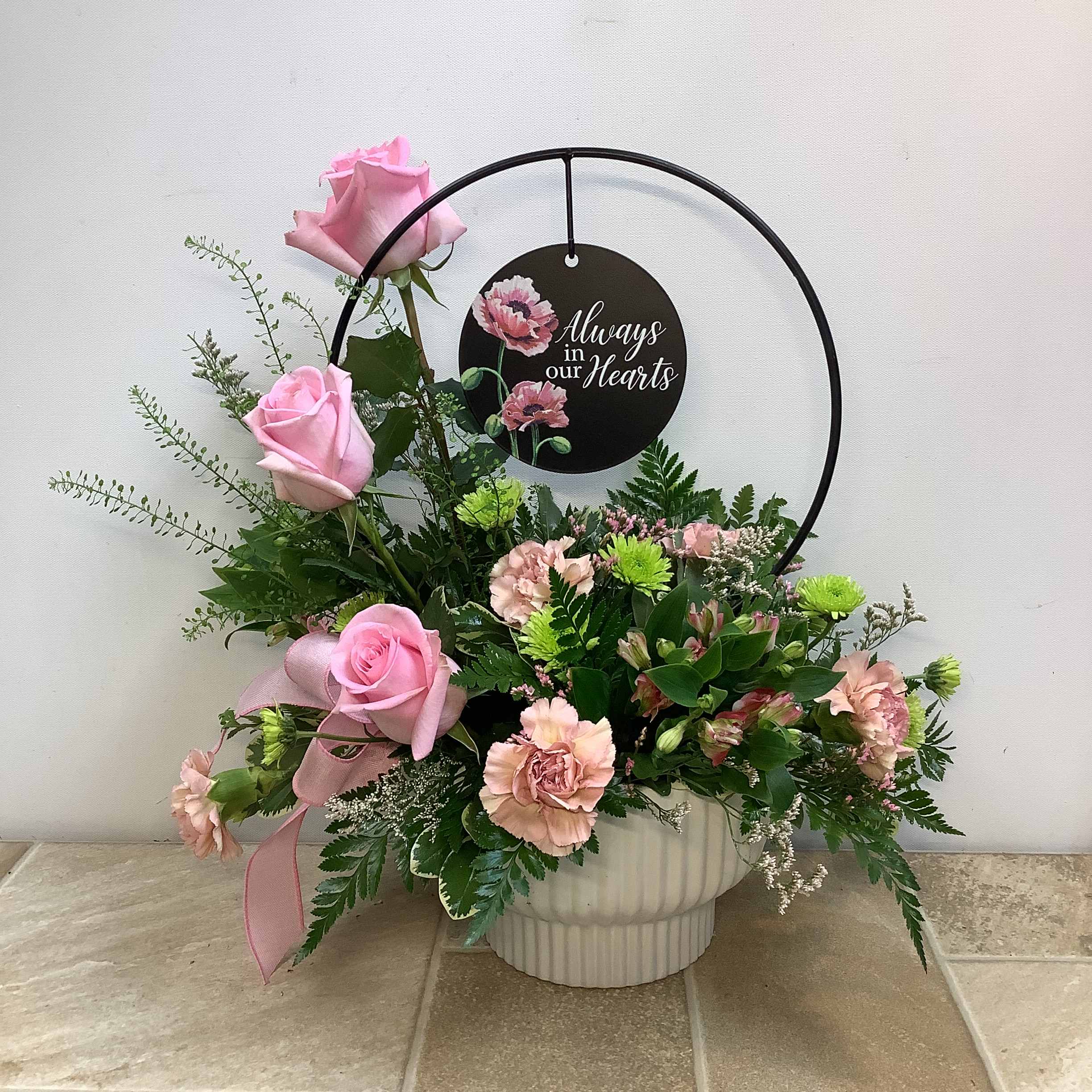 Beautiful Memories Beautiful Memories - "Beautiful Memories" is expertly arranged in a white ribbed ceramic container and includes a decorative metal hoop and disc with the phrase “Always in Our Hearts” for future placement in a garden or plant. Pink roses are prominently placed and symbolize admiration and gratitude, and soft pink carnations and charmellia represent remembrance and eternal love. Accent flowers and lush greenery add texture and volume. A delicate pink bow adds a personal touch. It measures 18” wide x 19” high. Primary flowers: rose, carnation, charmellia