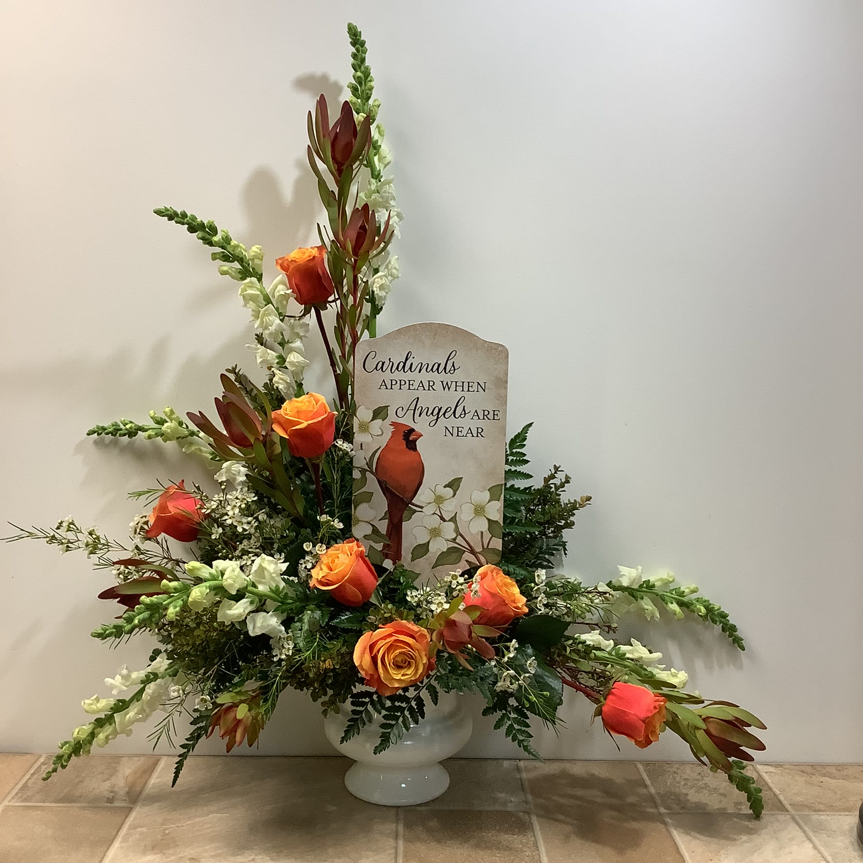Cardinals Appear Bouquet Cardinals Appear Bouquet - "Cardinals Appear" is a graceful and heartfelt tribute arrangement designed to offer comfort and remembrance. It is artfully presented in a classic white pedestal, urn-style vase. At the center is a beautiful garden stake with the words “Cardinals appear when angels are near”, that is sure to become a treasured keepsake. The flowers include orange-toned Cherry Brandy roses which add warmth and symbolize admiration and love, creamy white snapdragons to represent strength and grace, rusty red leucadendron for an earthy texture, and charming white accent flowers. Lush greenery enhances the arrangement's fullness. It measures approximately 30” wide x 36” tall. Primary flowers: Rose, snapdragon, waxflower
