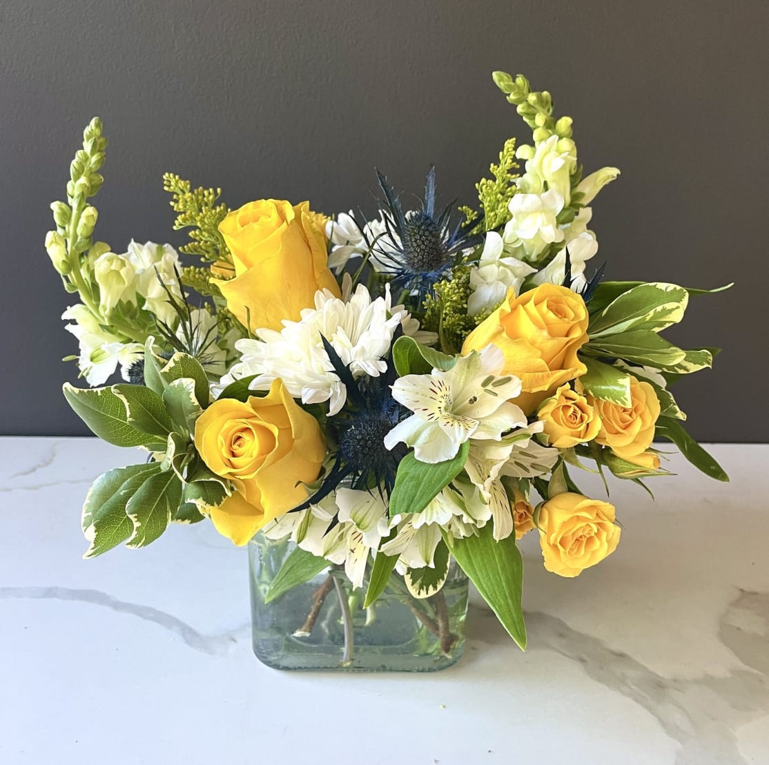 Golden Sunshine Elegance Bouquet - Lovely white and yellow flowers make up this dainty arrangement in a cube vase. Roses snapdragons and thistle are the main characters of this eccentric design. We reccomend this design for new babies (ask us about a pink version!), birthdays, and get well soon occasions.   *The types of flowers and greens used may vary depending on availability, however we will strive to maintain the colors and overall character of the arrangement as shown. 