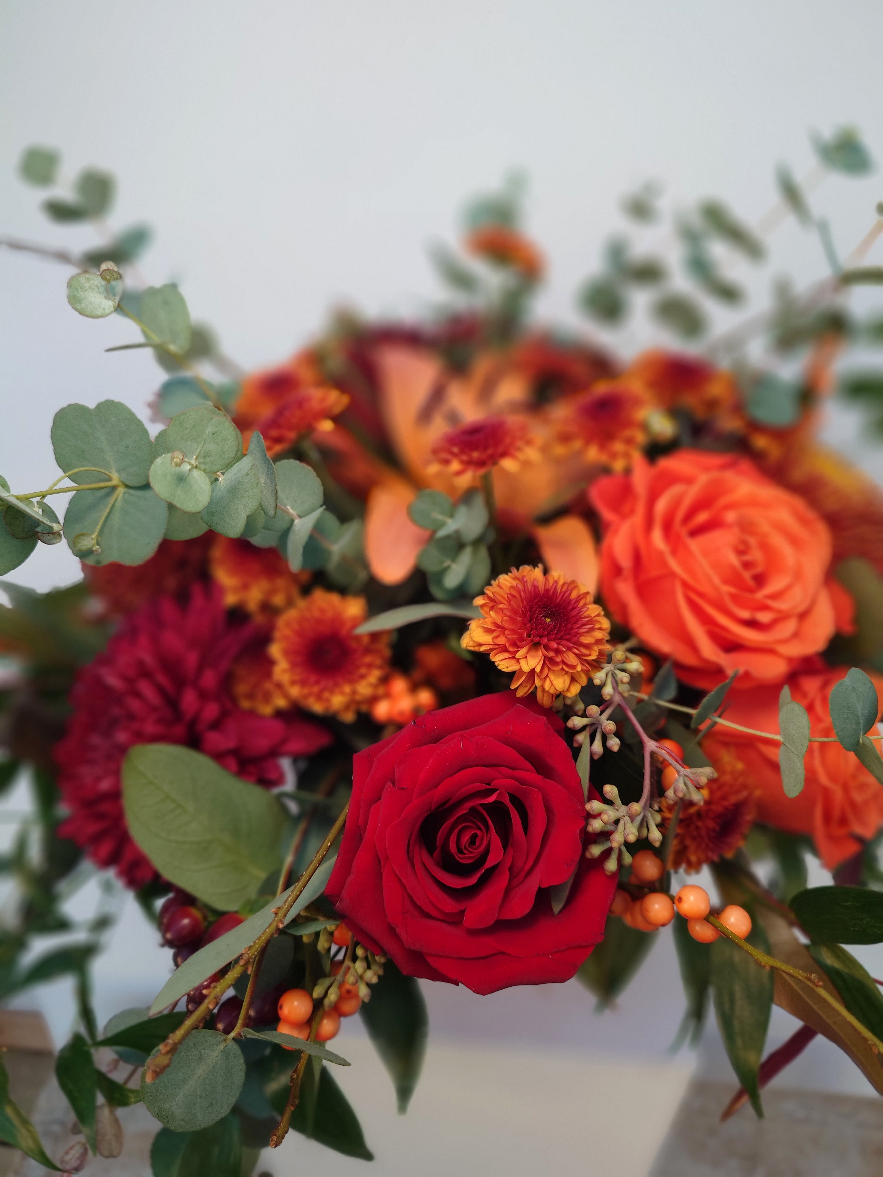 Cascadilla Cornucopia - Capture the cascading beauty of Cascadilla Falls with our &quot;Cascadilla Cornucopia&quot; centerpiece, designed to celebrate Thanksgiving with flair. This lush arrangement in a striking red metal vase features a bountiful mix of blooms and foliage that spill gracefully over the edges, reminiscent of the falls themselves.  Peach and burgundy winterberries add a splash of warm, seasonal color, while rich red roses, golden mums, and tall greens create texture and height. Cascading greenery flows beautifully, adding a sense of movement and natural elegance. Perfect for tables in Ithaca, Lansing, Trumansburg, and nearby areas, the &quot;Cascadilla Cornucopia&quot; evokes the natural wonder of the Finger Lakes region, bringing a touch of its splendor to your holiday table.