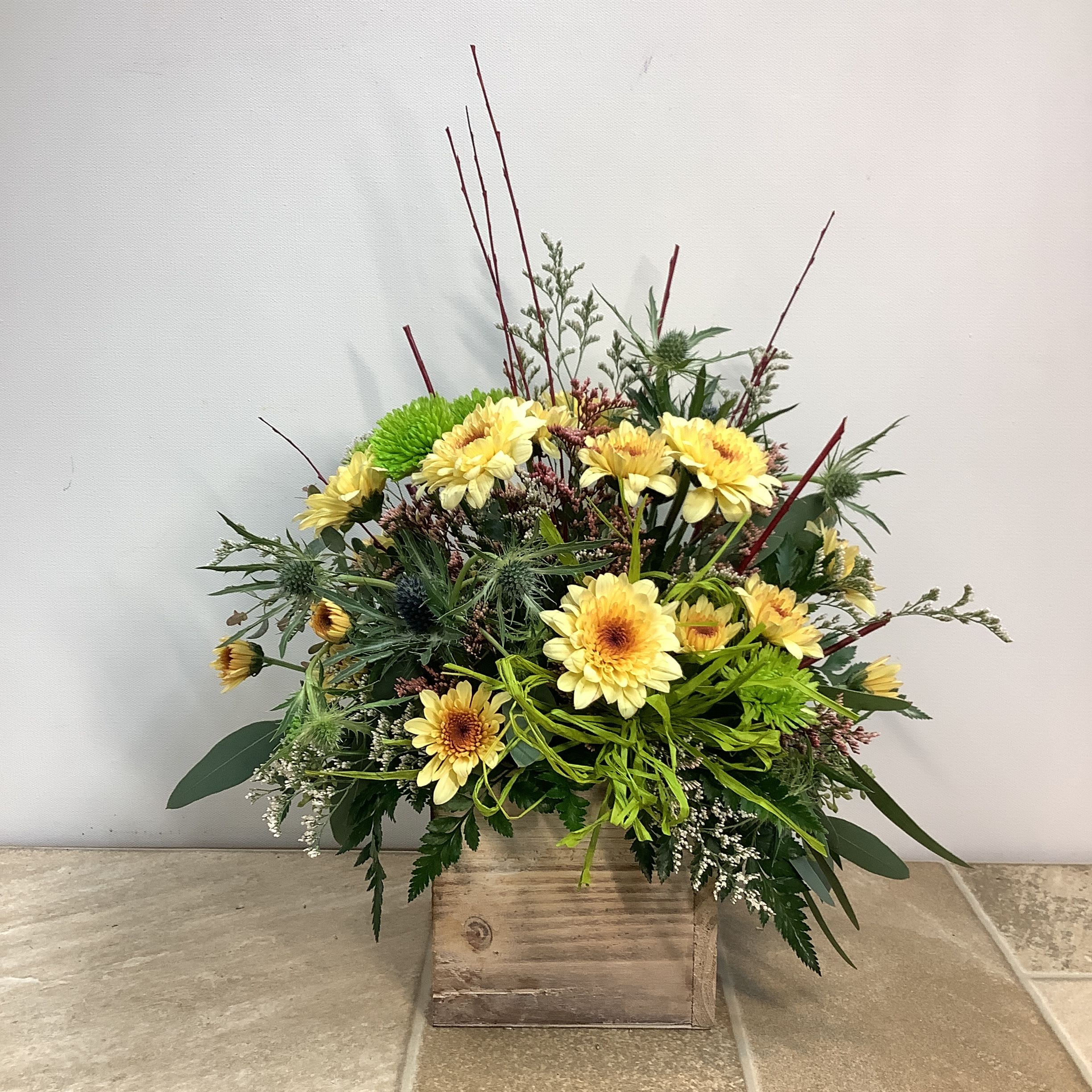 Crème Brûlée Crème Brûlée - Crème Brûlée is the name of the creamy yellow chrysanthemums used in this arrangement. Blue thistle, bright green mums, and different colored accent flowers, along with a variety of green foliage, add color and texture. The rustic wooden box that holds this wildflower-style bouquet adds a touch of country charm. Approximate size: 13” wide x 16” tall. Primary flowers: mum, chrysanthemum