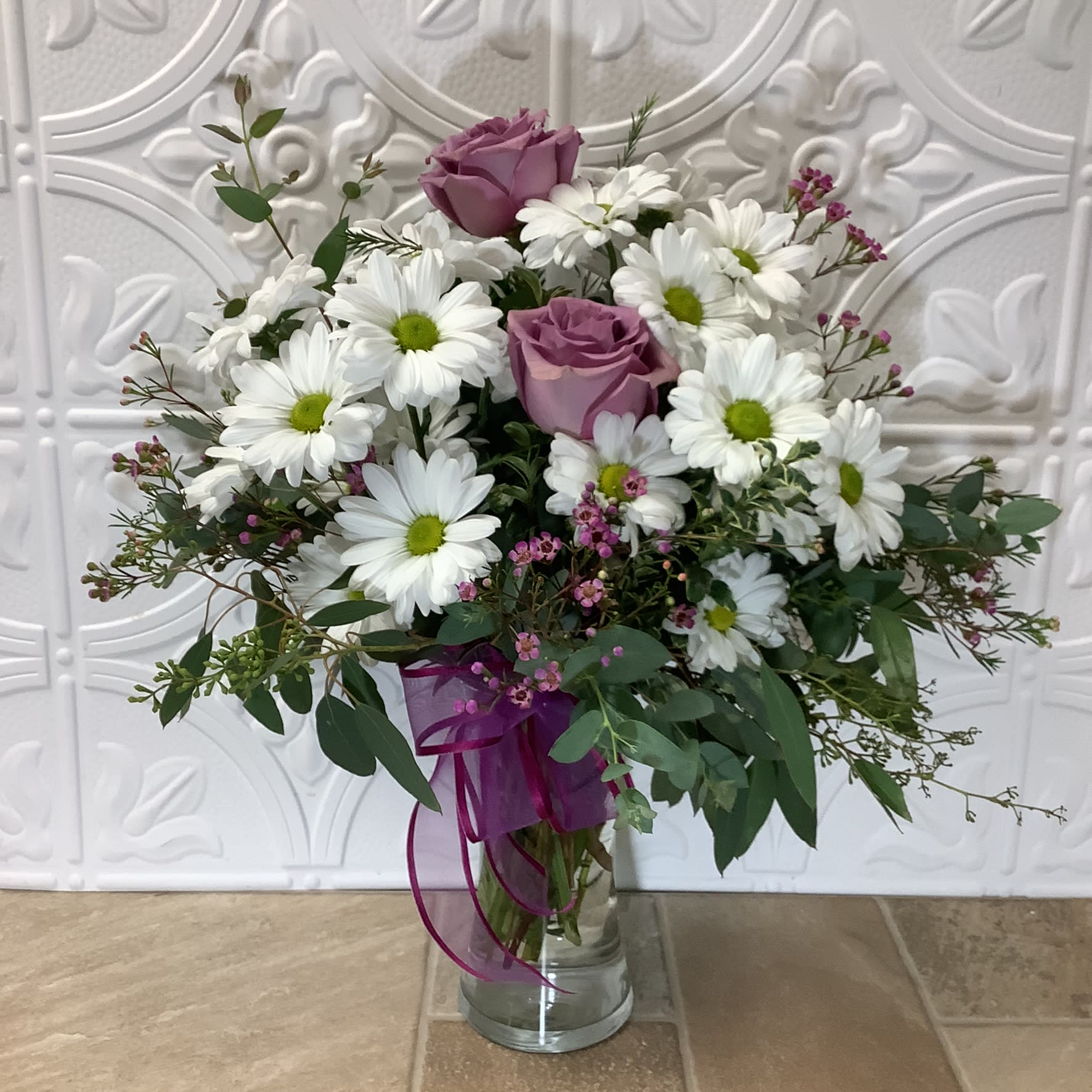Daisy Daisy - This floral arrangement is a beautiful and classic-looking bouquet featuring a mix of charming, white daisies and sweet, light purple/lavender roses. Delicate purple waxflowers, eucalyptus and other greens add lushness and texture to this harmonious design. The arrangement is presented in a clear glass vase with a purple ribbon tied around the neck. Primary flowers: daisy, rose waxflower