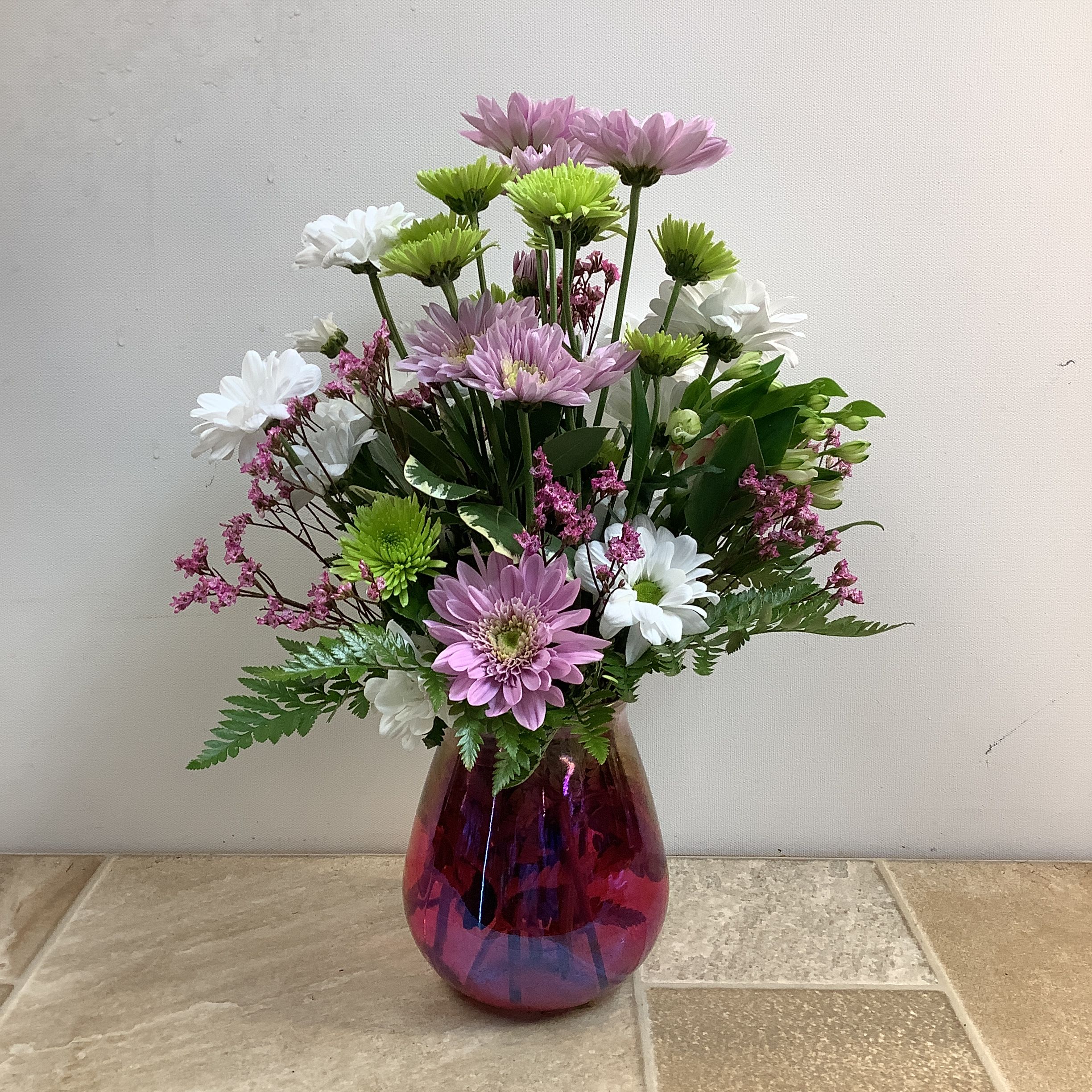 Daisy Delight - This vibrant floral arrangement, in a bright pink vase, includes sweet lavender and white daisies, surrounded by whimsical bright green mums, charmelia, and pink accents. Assorted green foliage adds depth and texture to this cheerful design.  Approximate size: 11” wide x 14” tall.  Primary flowers: daisy, mum