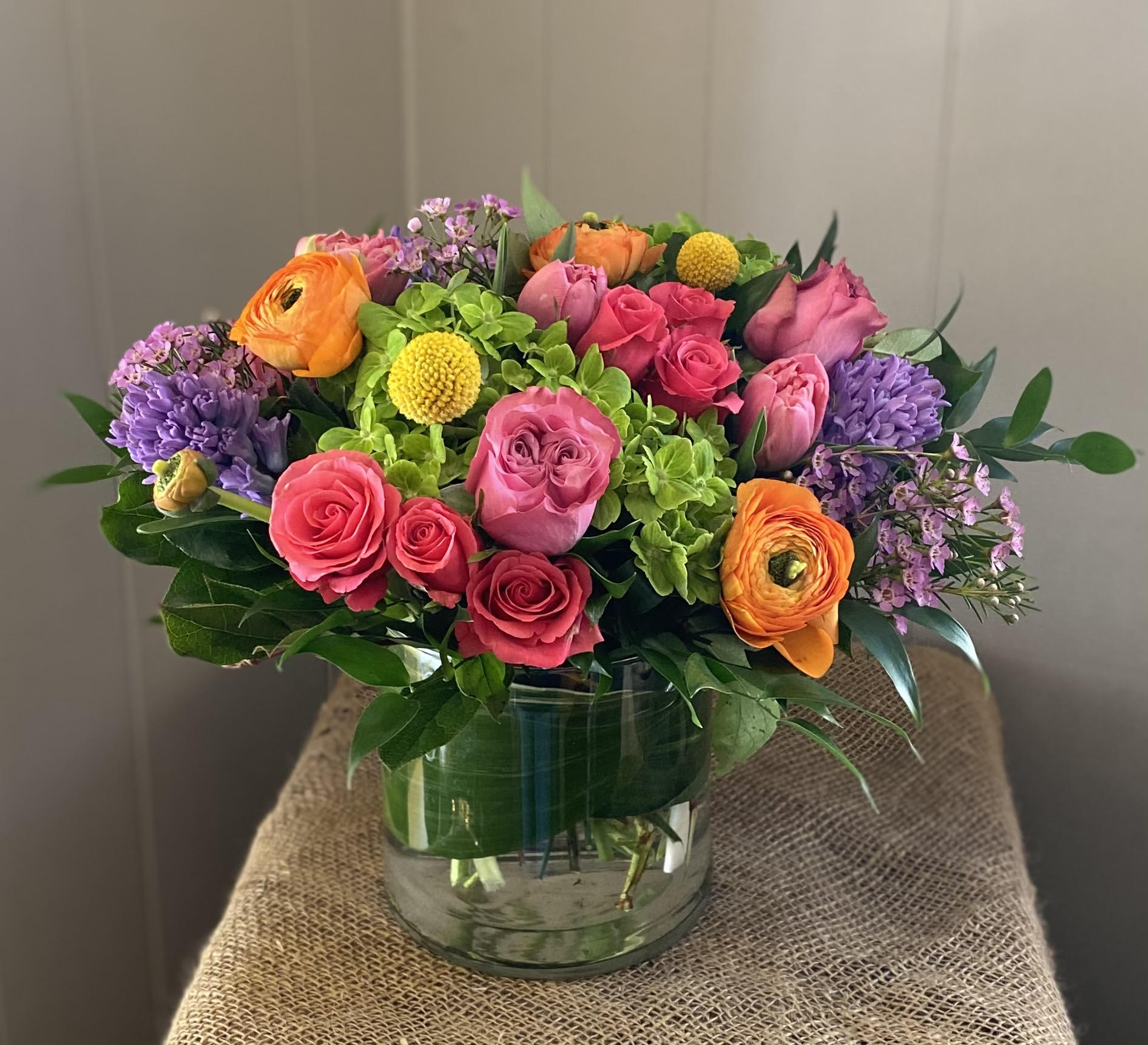 Rainbow Aura Rainbow Aura - If you need to brighten someone's day, there's no better way than with this arrangement! A stunning combination of green hydrangea, pink roses, orange ranunculus, purple hyacinth, and yellow craspedia, the Rainbow Aura always knocks it out of the park.