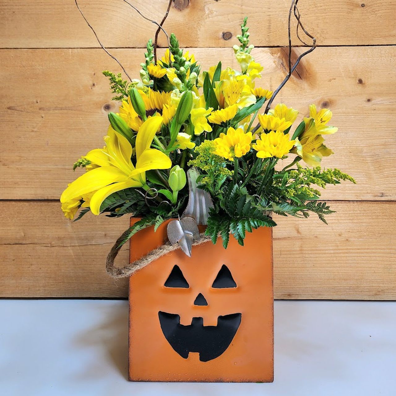 Jack's Golden Glow - Brighten the spooky season with a burst of cheerful charm! Jack’s Golden Glow is a radiant Halloween flower arrangement that pairs classic fall festivity with sunny warmth. This glowing bouquet features vibrant yellow snapdragons, lilies, alstroemeria, daisies, and bold sunflowers, all blooming like sunshine against the golden tones of autumn. Solidago adds texture, while whimsical curly willow branches rise like dancing shadows above it all. Displayed in a smiling jack-o'-lantern tin container, this piece is the perfect blend of Halloween spirit and seasonal cheer—ideal for welcoming guests or lighting up a festive table.  Approx. 22&quot; H x 10&quot; W