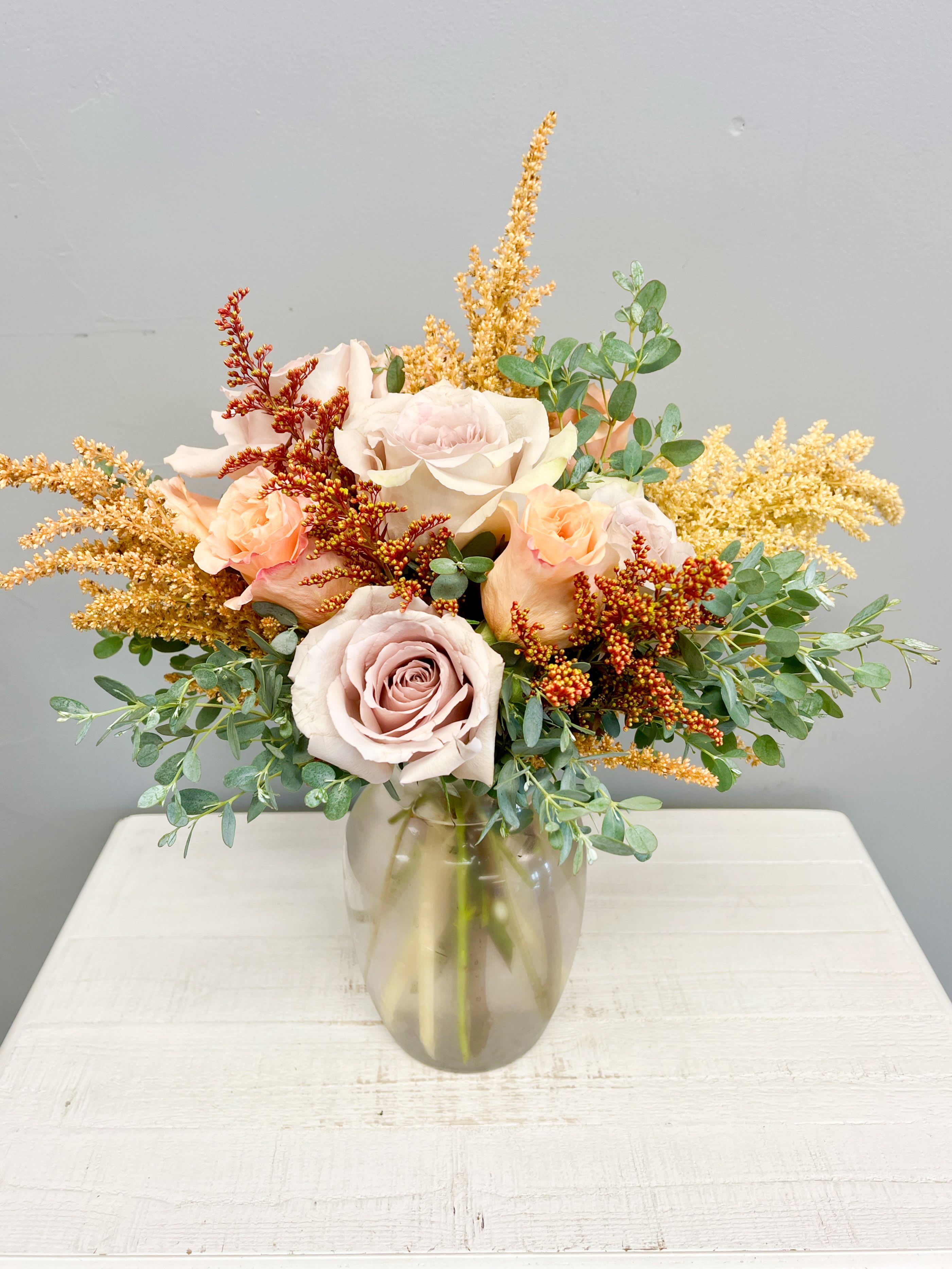 Amber Glow - Amber Glow Vase Arrangement Warm, radiant, and timeless—this arrangement captures the golden beauty of the season. Featuring premium roses in shades of champagne and blush, accented with soft peach spray roses, golden solidago, rust astilbe, and fresh eucalyptus, this bouquet is a perfect gift to brighten someone’s day or add elegance to any room.