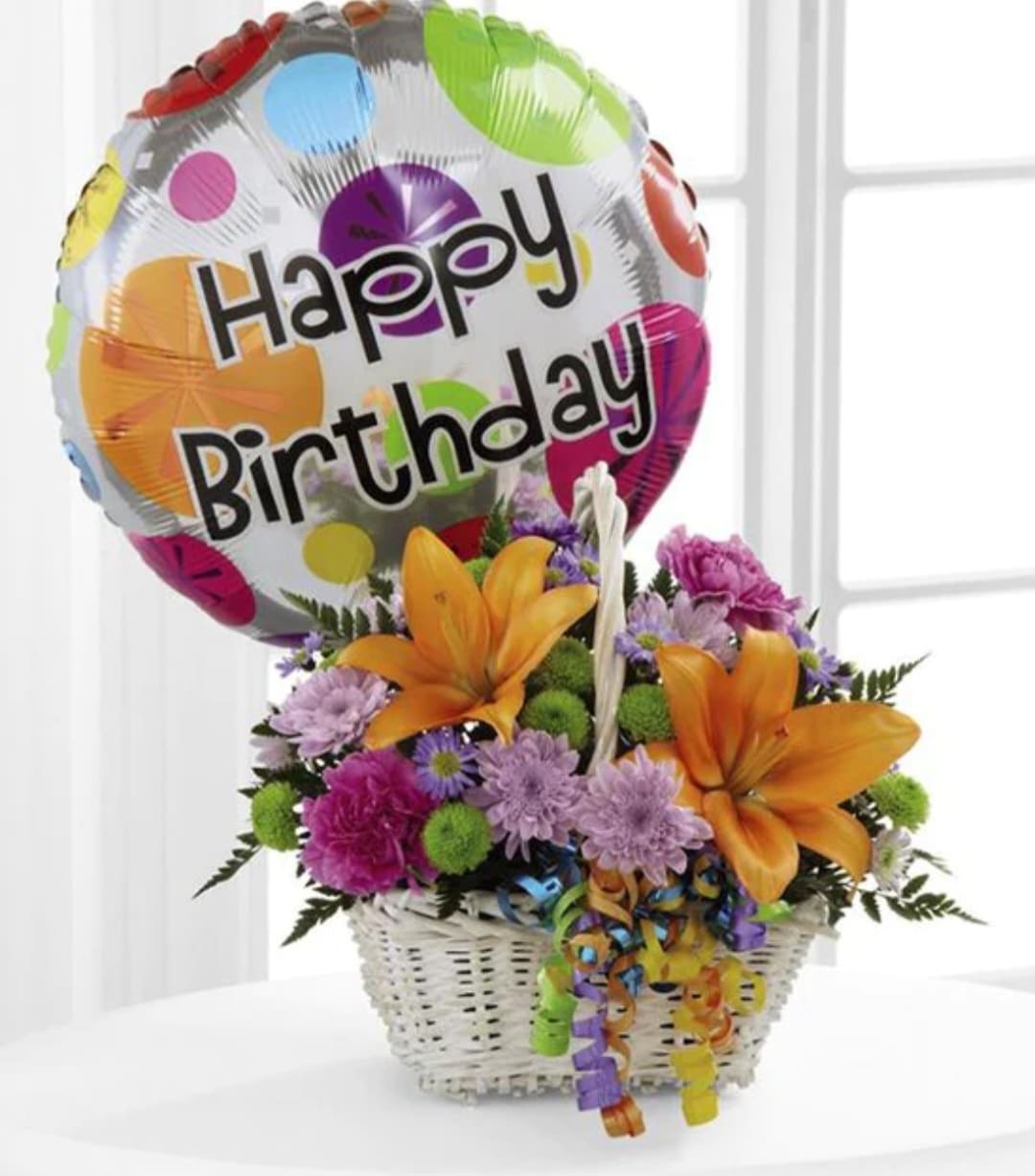 A birthday basket combo - A cute small birthday basket mixed with bright in season blooms carnations, chrysanthemums accompany by a birthday balloon.  The balloon does not float in the air it is filled and it is attached very close to floral arrangement it is an accent piece to floral arrangement. The basket may be white or brown depending on inventory  