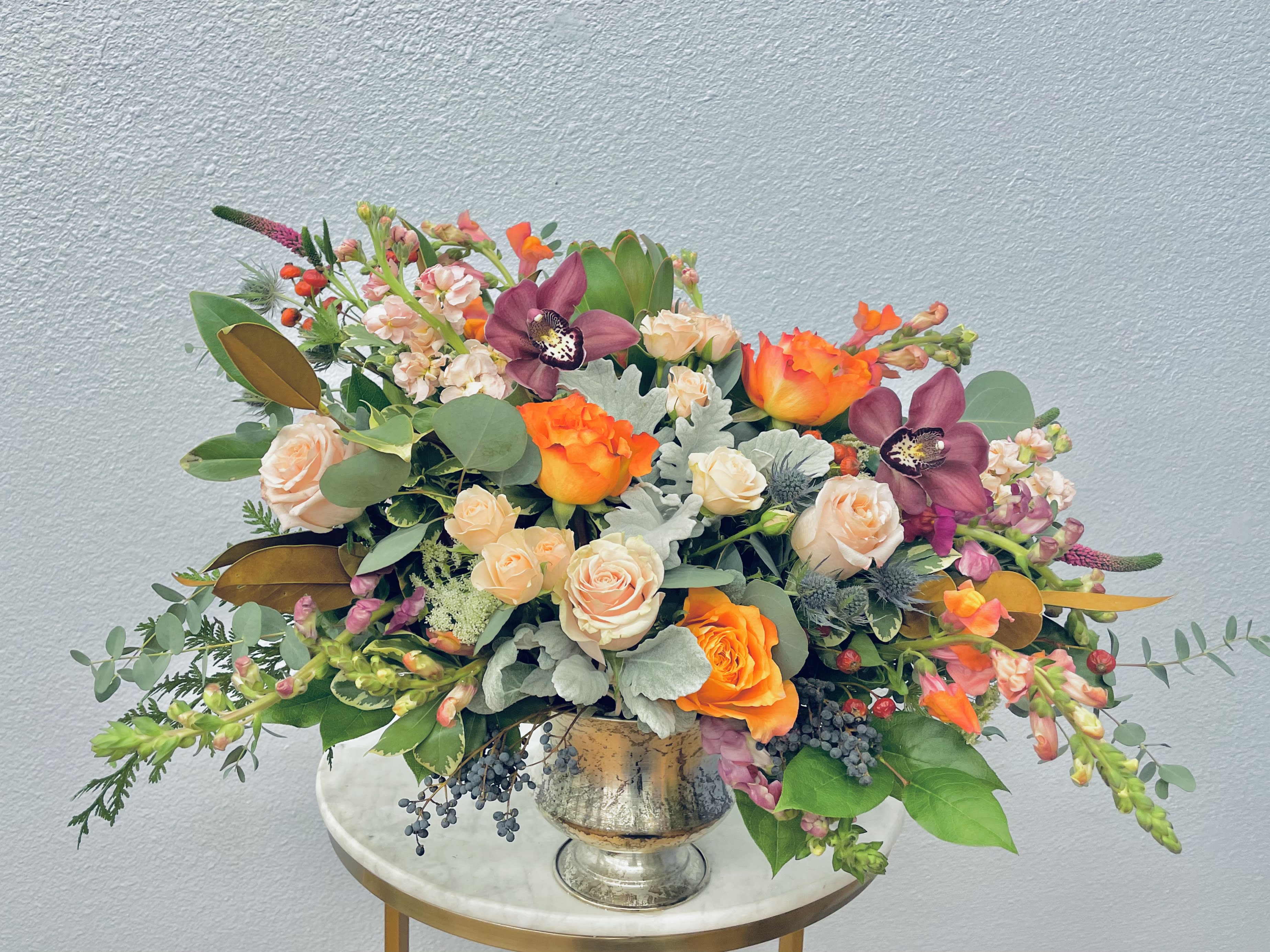 Genevieve - Color Palette: blush, apricot, purple, pink, green Blush and apricot roses paired with salmon snapdragons, pastel spray roses, veronica and burgundy cymbidiums tucked into botanical eucalyptus and seasonal greens in a simple silver vase.   *Photo shows deluxe upgrade*