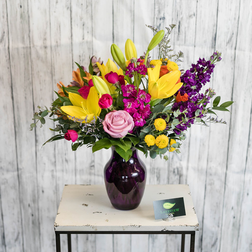 Parade - A delightfully vibrant mix of blooms that are presented in a purple, glass vase.  Approximate Dimensions: 20&quot;H x 20&quot;W