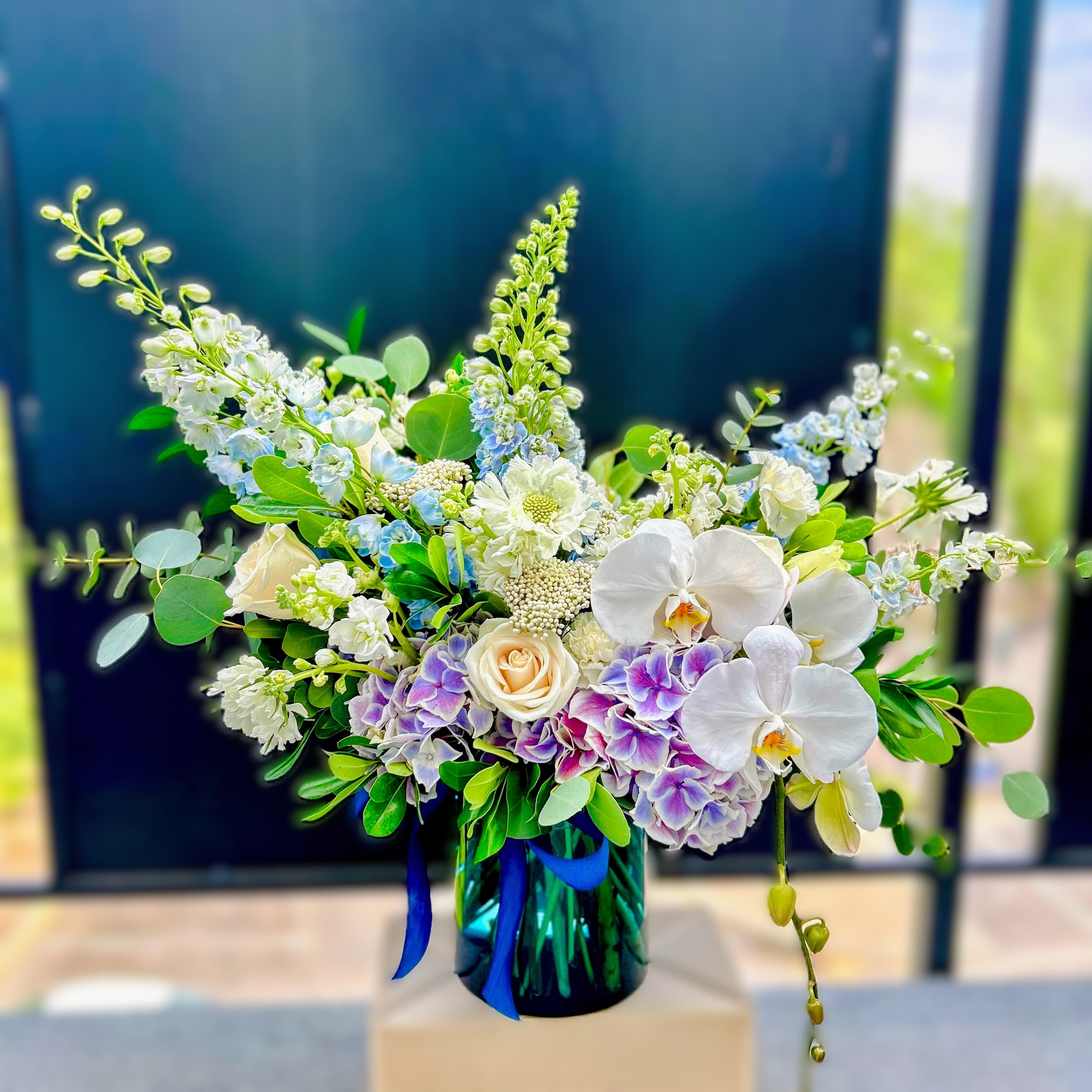 Partly Cloudy - Like a sky after rain, Partly Cloudy drifts between softness and light. Cool blues, creamy whites, and violet tones gather in a lush cloud of blooms, accented with airy textures and elegant orchids. A moment of calm and wonder, captured in flowers.