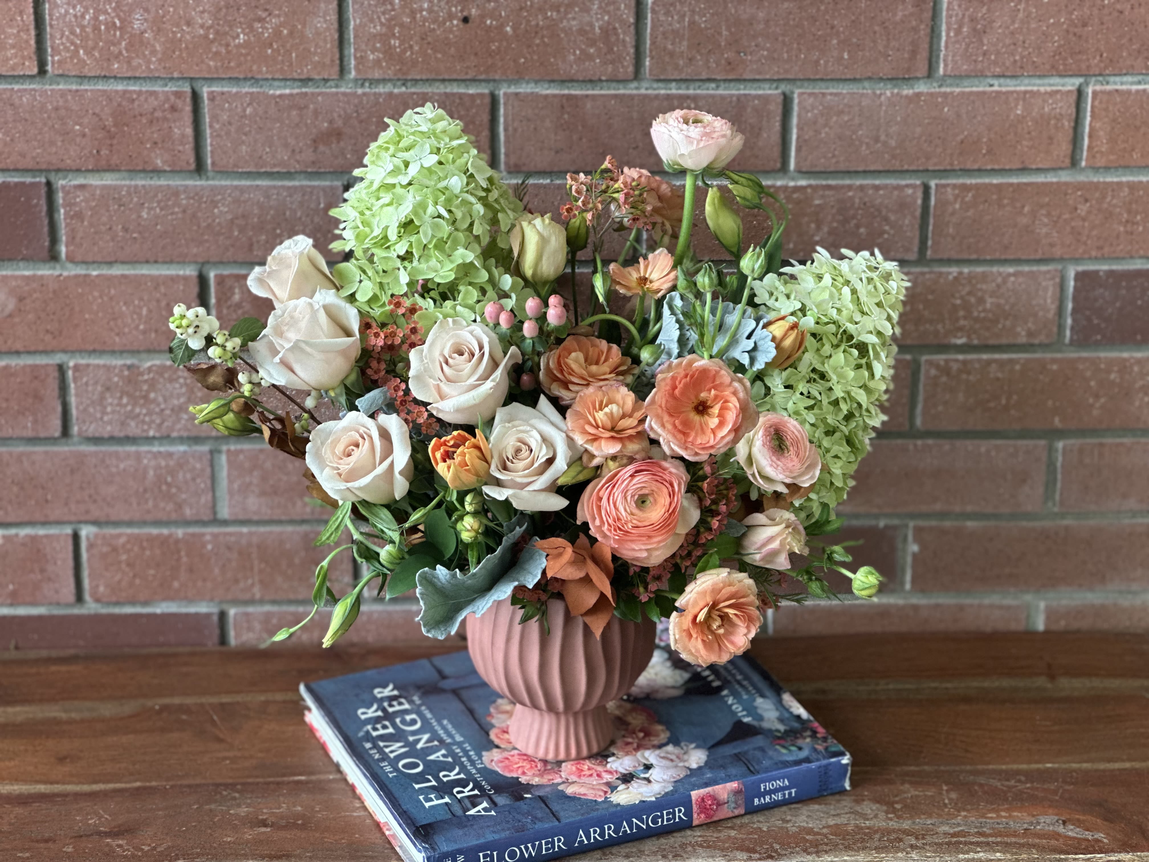 Sawgrass Serenade  - This enchanting arrangement sings with the soft, melodic tones of a Florida autumn evening. The coral and peach ranunculus echo the warm sunset hues that dance across the Everglades’ endless sawgrass, while the champagne roses mirror the golden light filtering through cypress canopies. The lime green hydrangeas celebrate Florida’s year-round vitality - like the persistent green of sawgrass that sways in gentle autumn breezes. Silvery dusty miller adds whispered texture reminiscent of Spanish moss and morning mist over the wetlands, while burgundy accents ground the composition like the deep, rich waters of the Everglades at twilight. Arranged in a fluted ceramic wave handmade vase. 