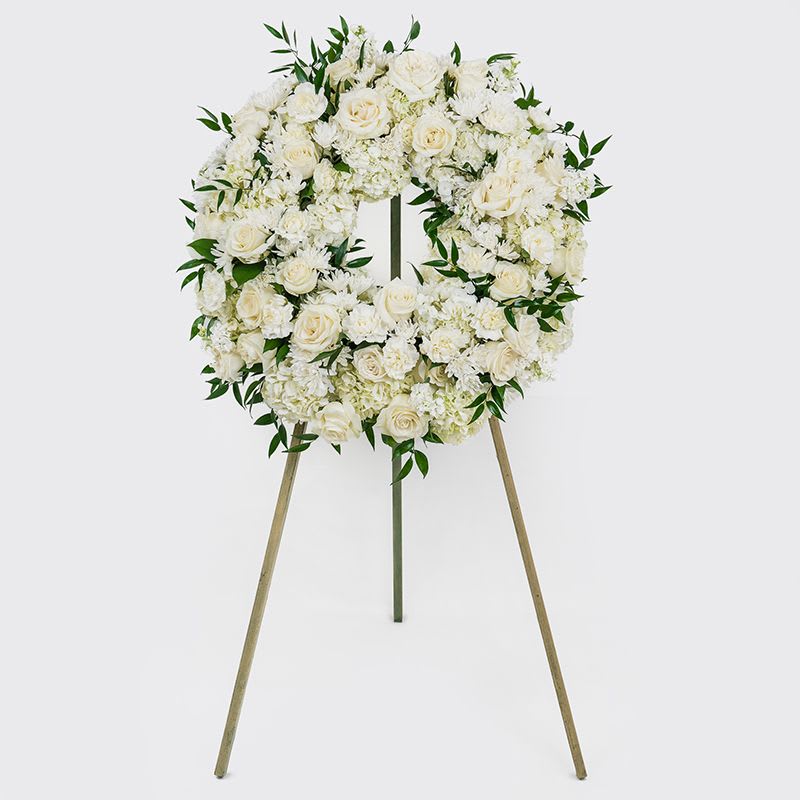 Funeral Wreath - A classic floral wreath featuring a mix of premium and seasonal white blooms accented by textural greenery. Designed with a timeless and structured yet natural feel. 18&quot; Wreath, Placed upon a 5’ wood easel, included.