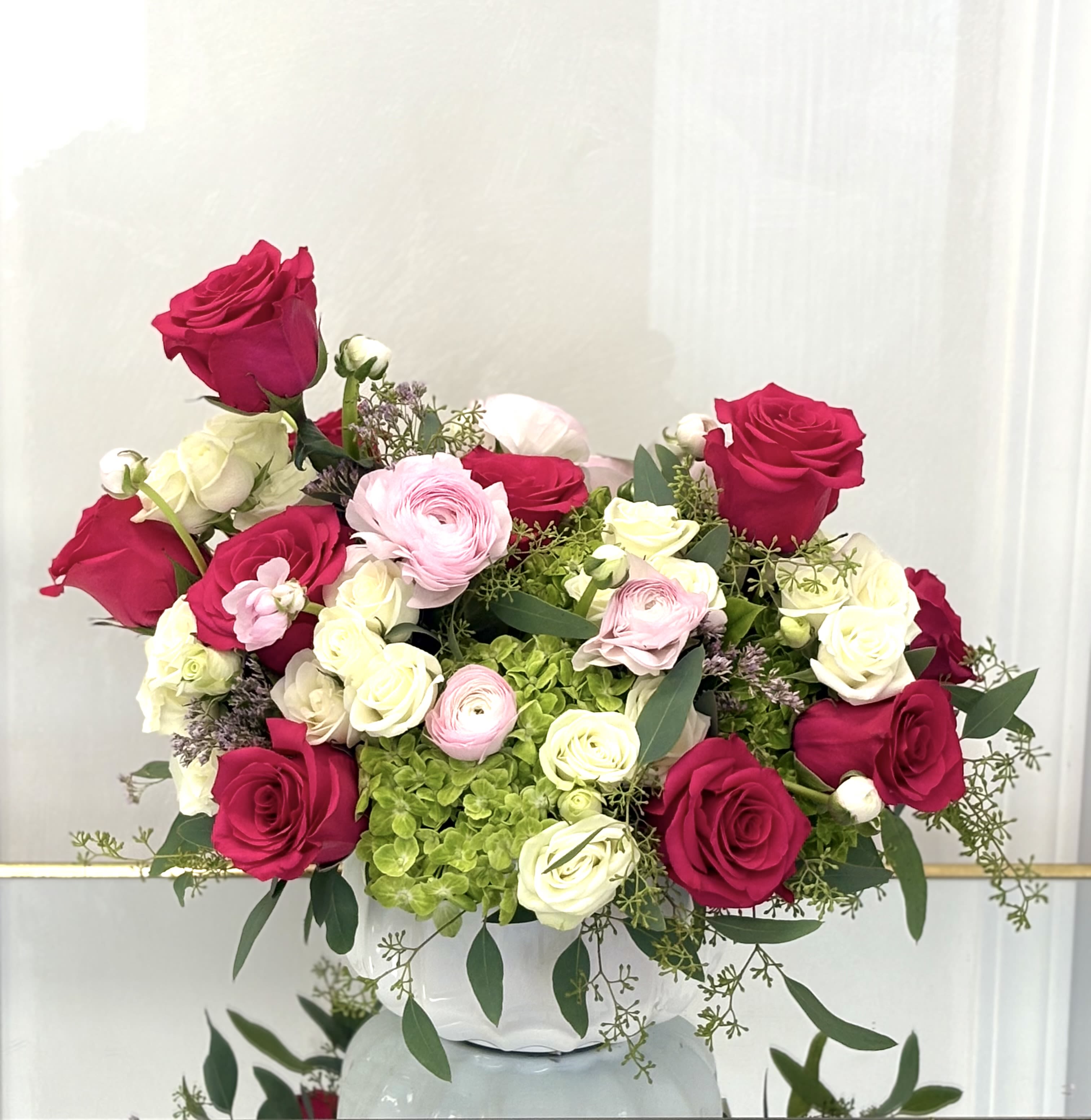 Blushing Grace By VLM Flowers -  Soft pink ranunculus and creamy roses dance between bold pink blooms, creating a romantic contrast of strength and sweetness. Nestled in a porcelain-white vase and accented with green hydrangeas and eucalyptus, this arrangement speaks the language of love with a timeless, graceful tone.  Perfect For: Anniversaries, birthdays, or expressing admiration and elegance.