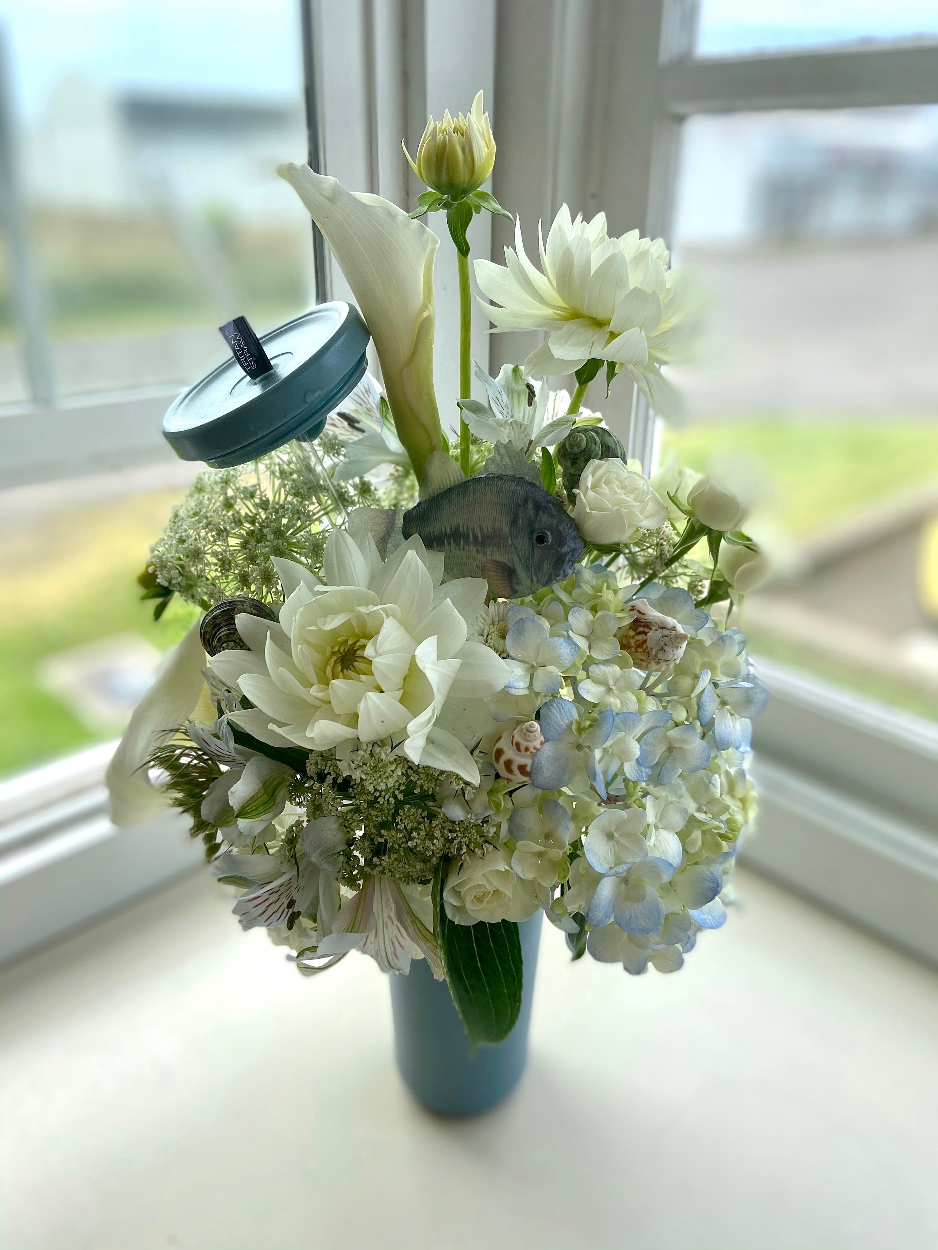 Seascape Tumbler Seascape Tumbler - This unique design is truly a gift that keeps on giving; enjoy fresh, ocean-themed flowers, and then an eco-friendly stainless steal container! This soft blue color pallet including hydrangea, dahlias, and calla lilies, is adorn with seashells and even a fish nestled in the center! This is perfect for an ocean loving recipient!