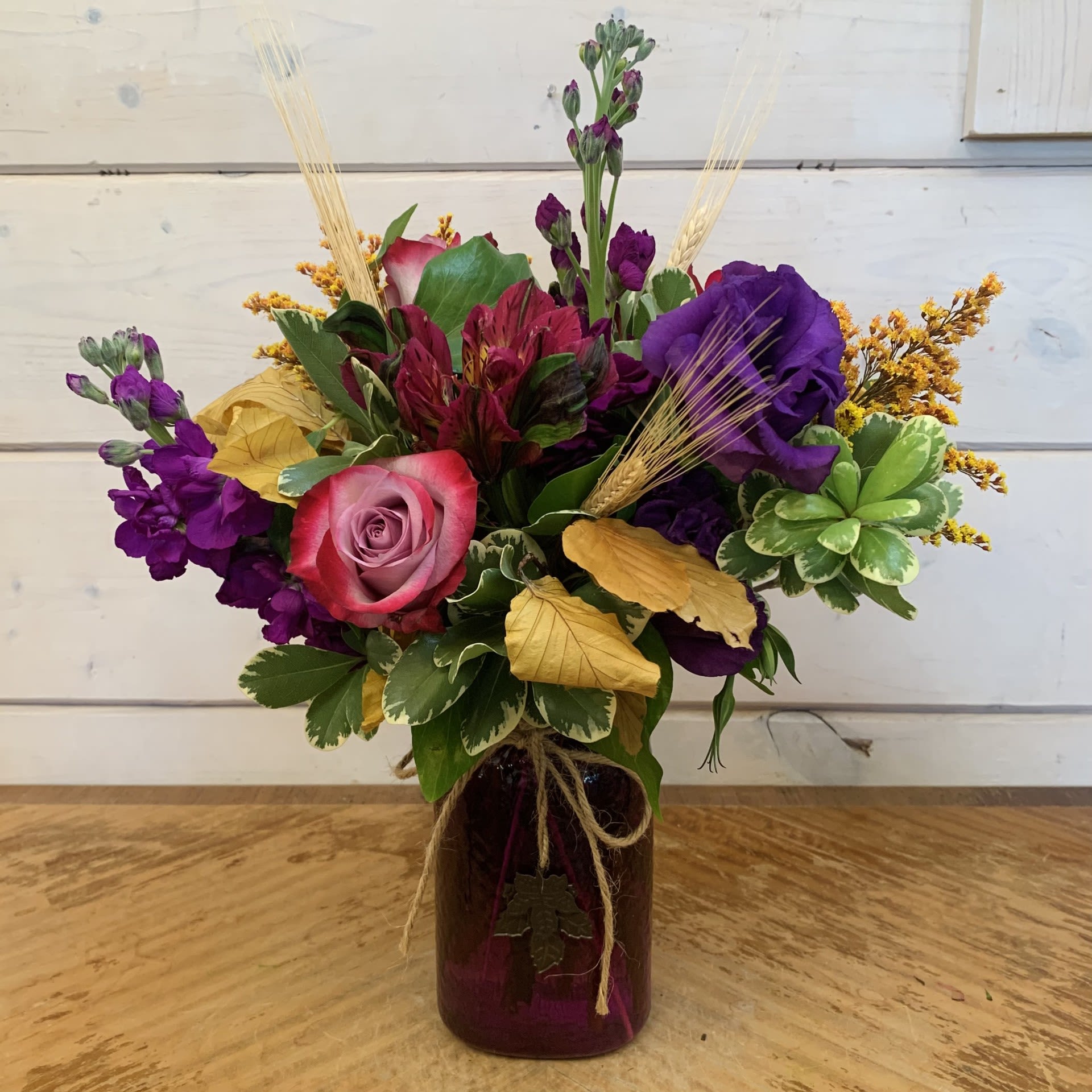 Autumn Loves Purple Autumn Loves Purple - Lavender roses, purple lisianthus, purple stock and purple alstroemeria with fall solidago, copper beech, and wheat in a textured purple glass jar with maple leaf charm. Approximately 13" tall and 10" wide