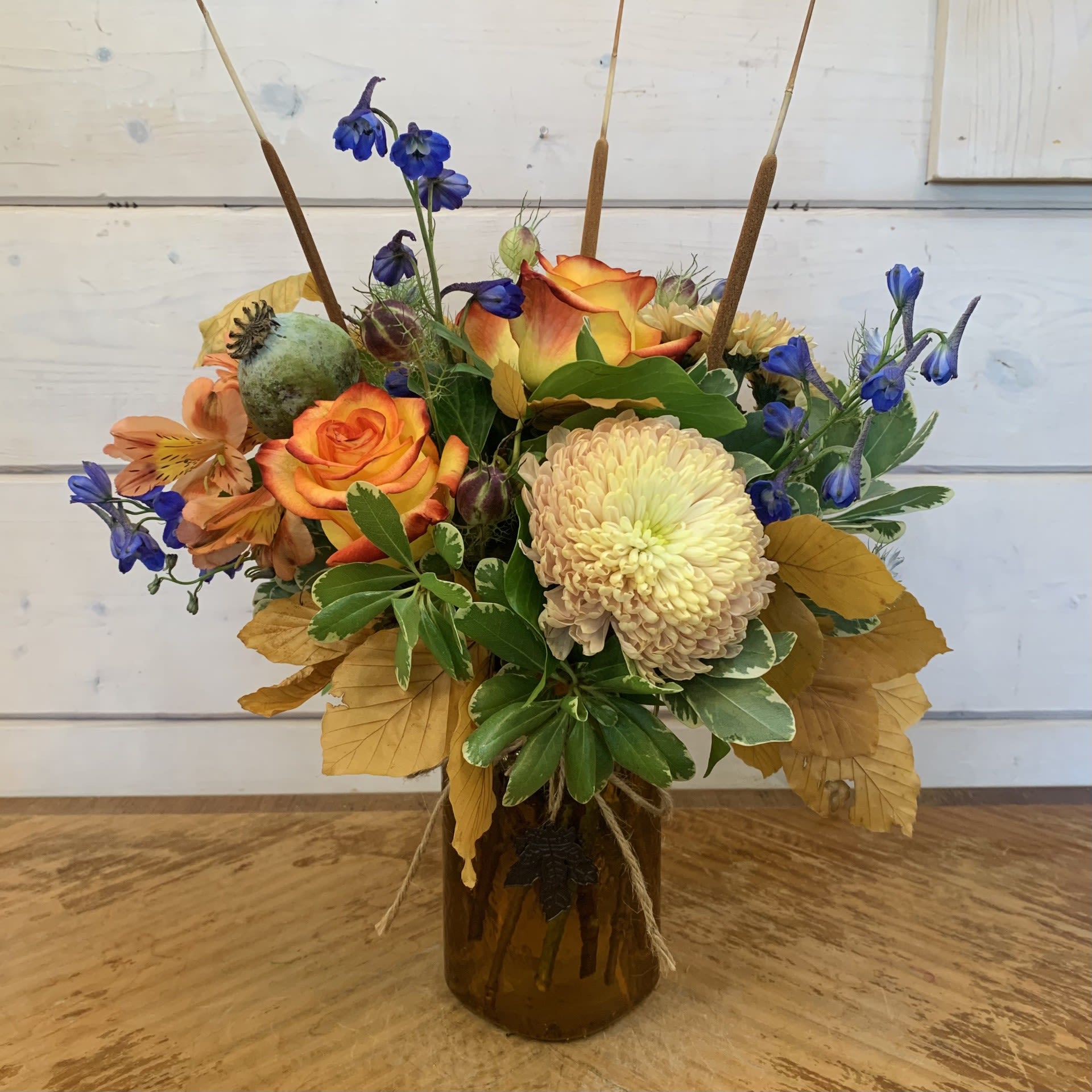 Fall Colors Fall Colors - Orange roses, blue belladonna, linnett cremon, fall crysanthemums, orange alstroemeria, with a poppy seed pod, nigella seed pods, and nigella blooms, copper beech, and cattails in a textured yellow mason with maple leaf charms. Approximately 13" high and 10" wide.