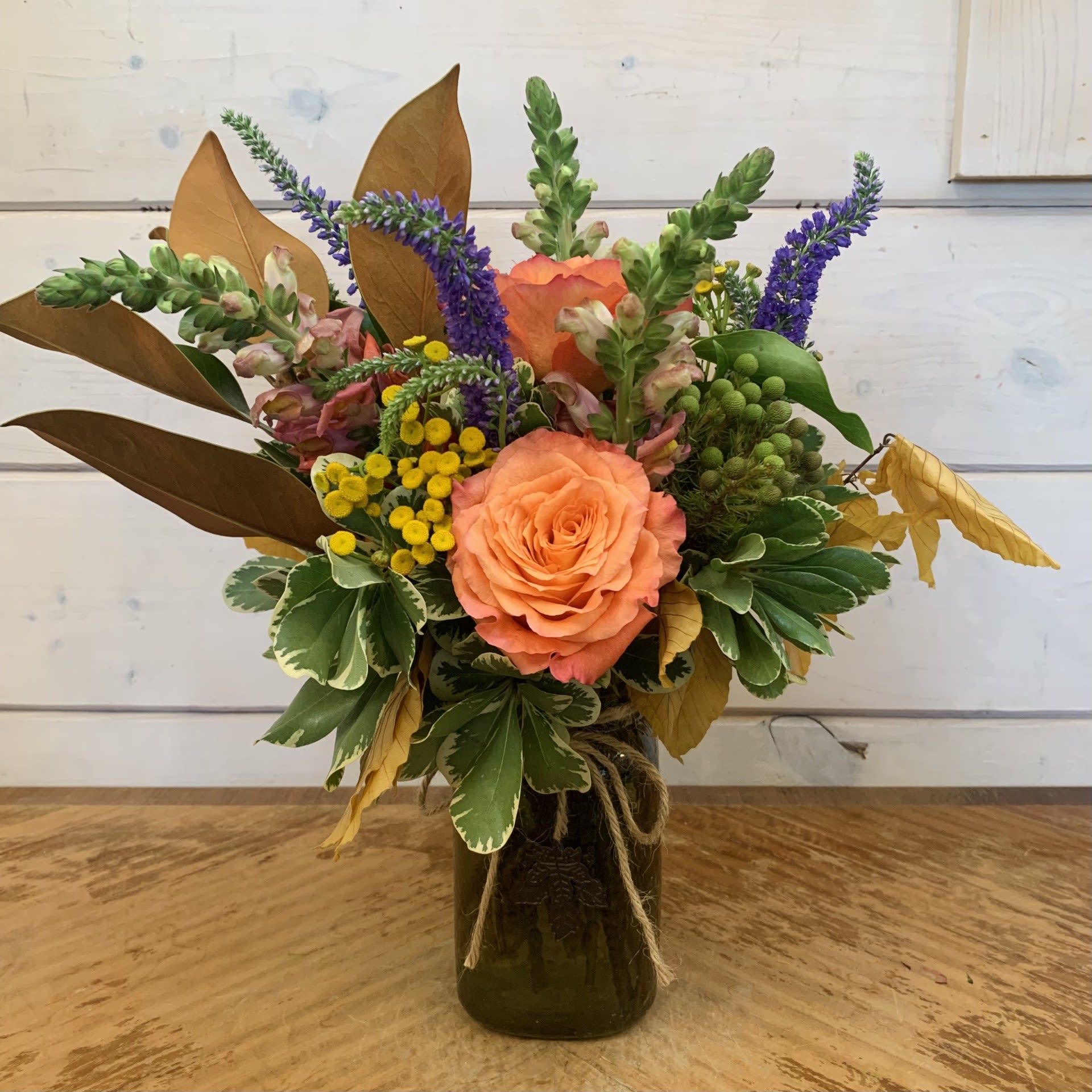 I Love Fall I Love Fall - Orange roses, purple veronica, orange snapdragons, brunia berries, and yarrow with copper beech and a magnolia leaf spray in a textured green mason jar with a maple leaf charm. Approximately 13" high and 10" wide.