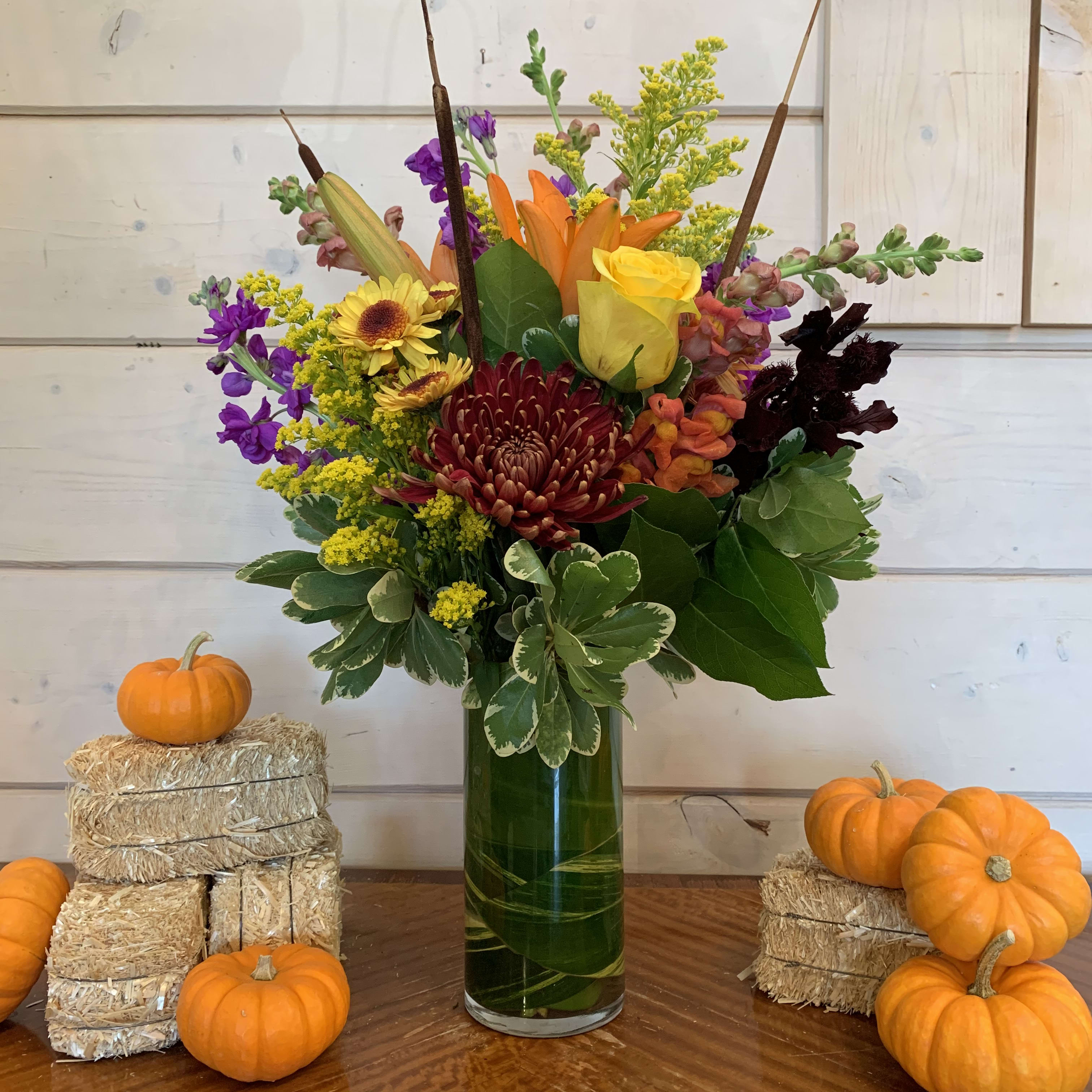 Shades of Autumn Shades of Autumn - Yellow roses, Orange lily, purple stock, orange snapdragons and fall chrysanthemums arranged in a leaf lined 9in cylinder with solidago and seasonal fall foliage. Approximately 18in tall and 12in wide