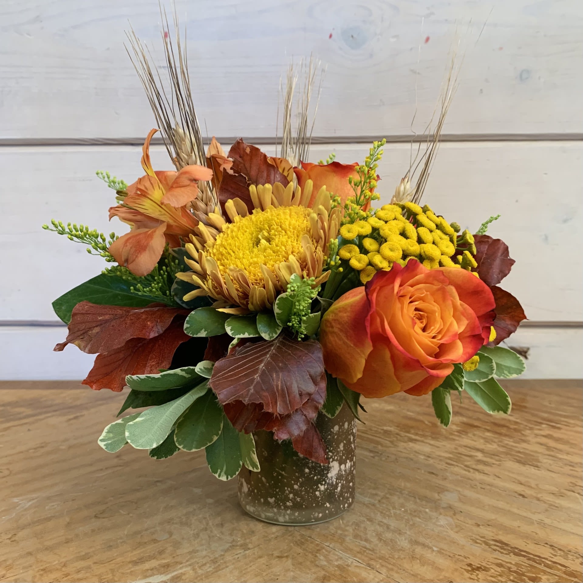 Wild Harvest - Orange roses, a gold cremon, orange alstroemeria, and yarrow in a peach frosted glass vase with seasonal filler flower, black wheat, and copper beech. Approximately 7in tall and 7in wide.