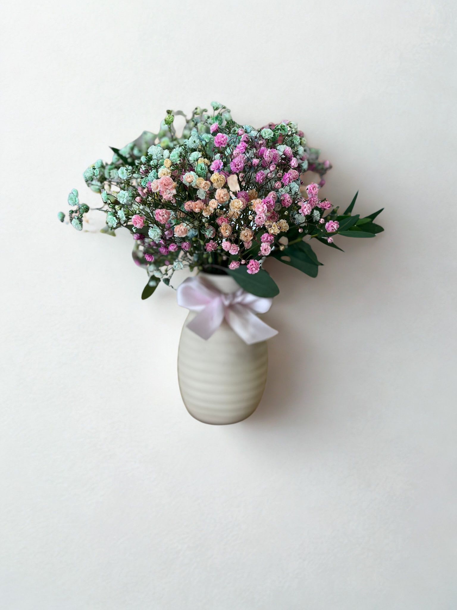 Brisa pastel-Pastel breeze - A delicate arrangement of gypsophila in pastel shades (pink, blue, lilac, and peach) presented in a white ceramic vase with a satin bow. Its romantic and elegant style makes it the ideal gift to express tenderness, love, and gratitude. Accompanied by a wax-sealed envelope, this arrangement is perfect for birthdays, thank-yous, or simply to brighten any space with a touch of sweetness.   Un delicado arreglo de gipsófila en tonos pastel (rosa, azul, lila y durazno) presentado en un florero cerámico blanco con lazo de satén. Su estilo romántico y elegante lo convierte en el detalle ideal para expresar ternura, amor y gratitud. Acompañado de un sobre sellado con cera, este arreglo es perfecto para regalar en cumpleaños, agradecimientos o simplemente para iluminar cualquier espacio con un toque de dulzura.