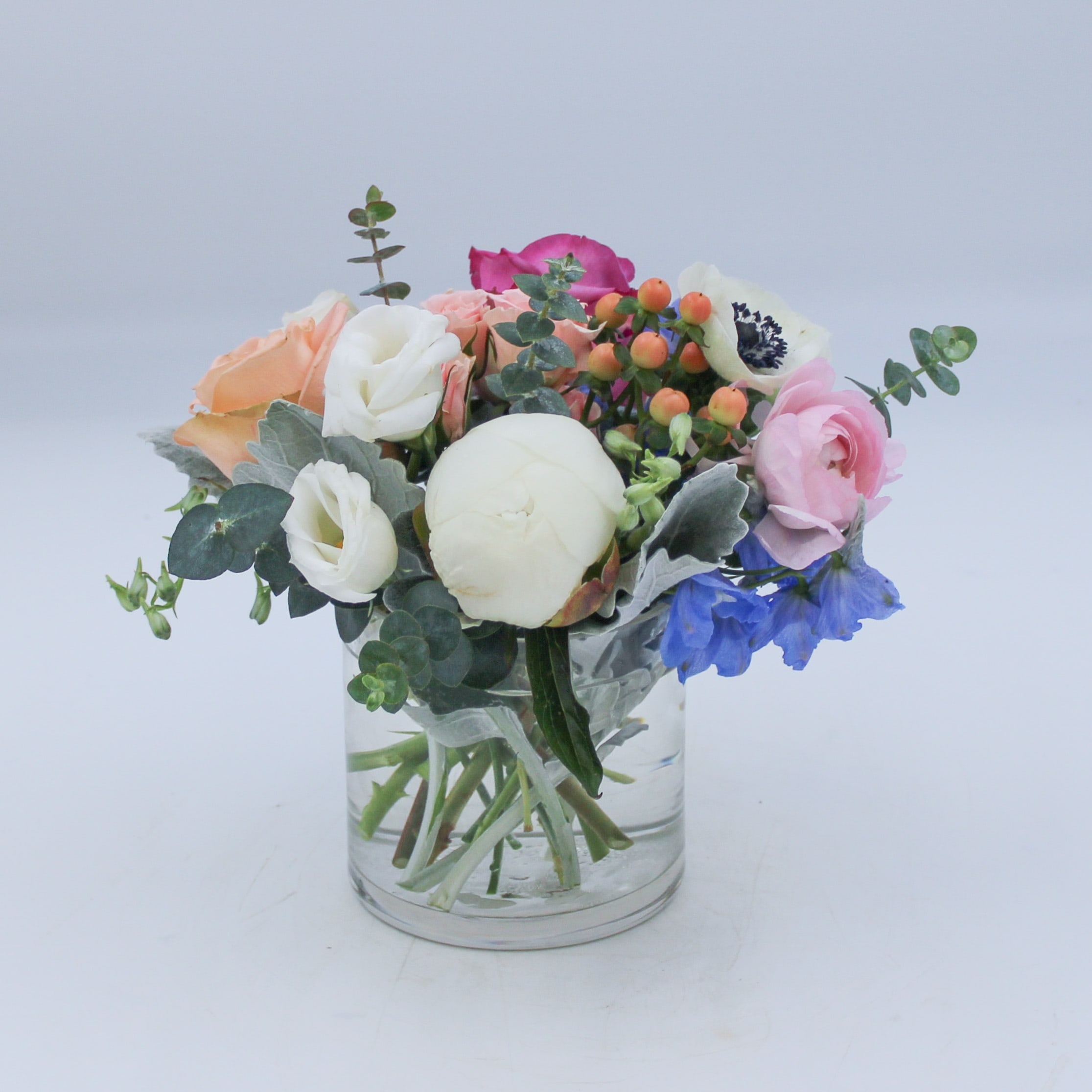 Petite Pastel - Petite Pastel is a charming arrangement of a White Peony (when in season), Deep Purple Rose, Peach Rose, White Anemone, Pink Ranunculus, Blue Delphinium, Peach Hypericum, Pink Spray Rose, Eucalyptus and mixed Ruscus. Petite Pastel is arranged in a 4 inch Cylinder and perfect for when you want to just send a little something luxurious.