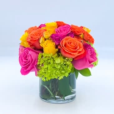 Tequila Sunrise - A vibrant display of Yellow, Hot Pink and Orange Roses and Spray Roses and Green Mini Hydrangea in a monstera leaf lined glass cylinder. Tequila Sunrise is arranged in a 5inch clear glass vase.