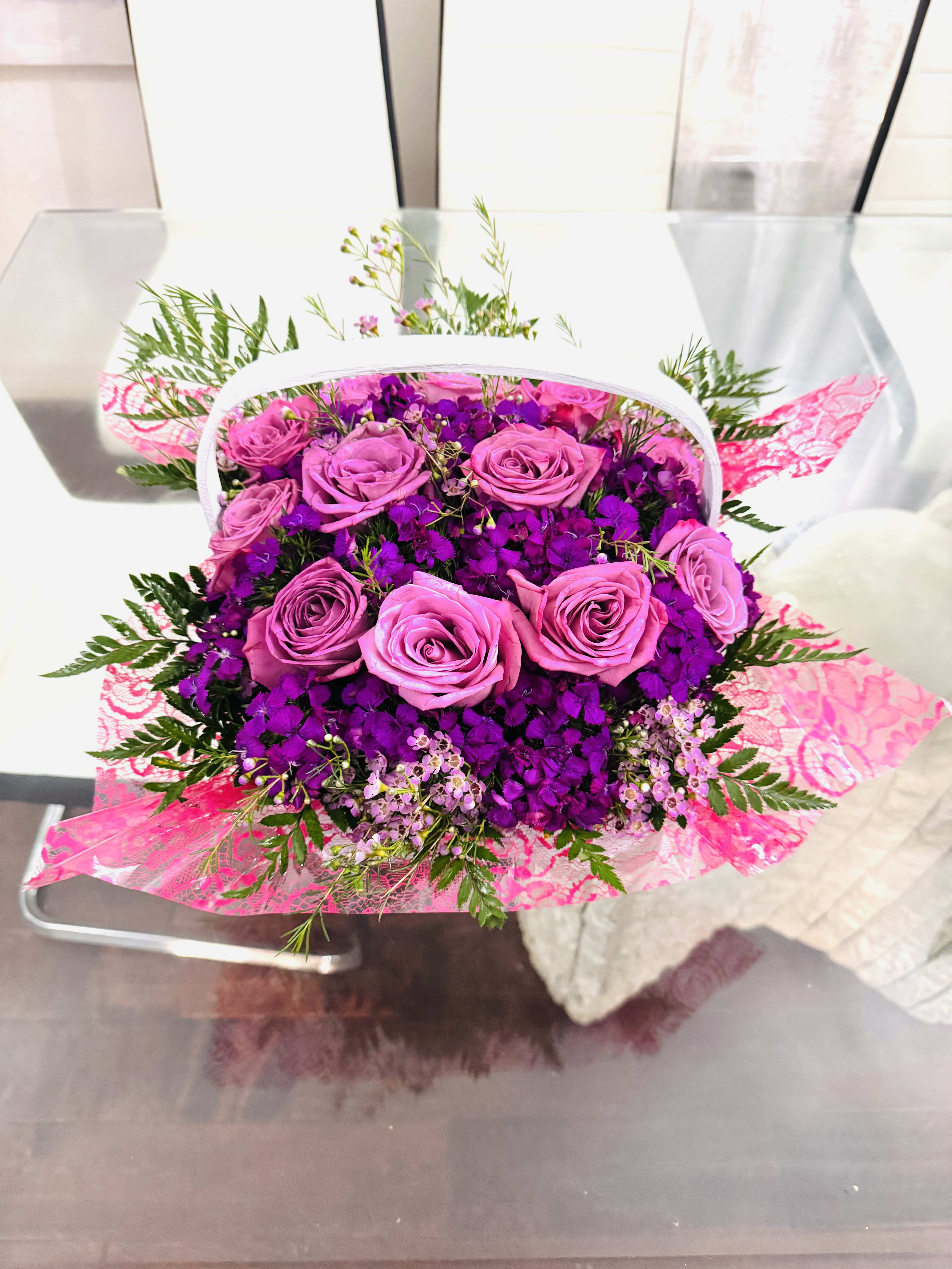 Purple Lovely Basket - This bouquet is an enchanting composition that radiates both romance and sophistication. It holds a graceful cluster of soft purple roses harmoniously paired with lush violet blooms, creating a symphony of purples that feels both vibrant and soothing. The touches of delicate wax flowers and airy greenery add refinement, while the pink lace accents lend a playful, feminine charm. It is a bouquet that speaks the language of elegance—gentle yet striking, refined yet full of warmth. Perfectly balanced between structure and softness, it evokes the feeling of a tender embrace or a whispered declaration of affection. This arrangement would not only brighten a room but also leave an impression of grace, beauty, and timeless love.