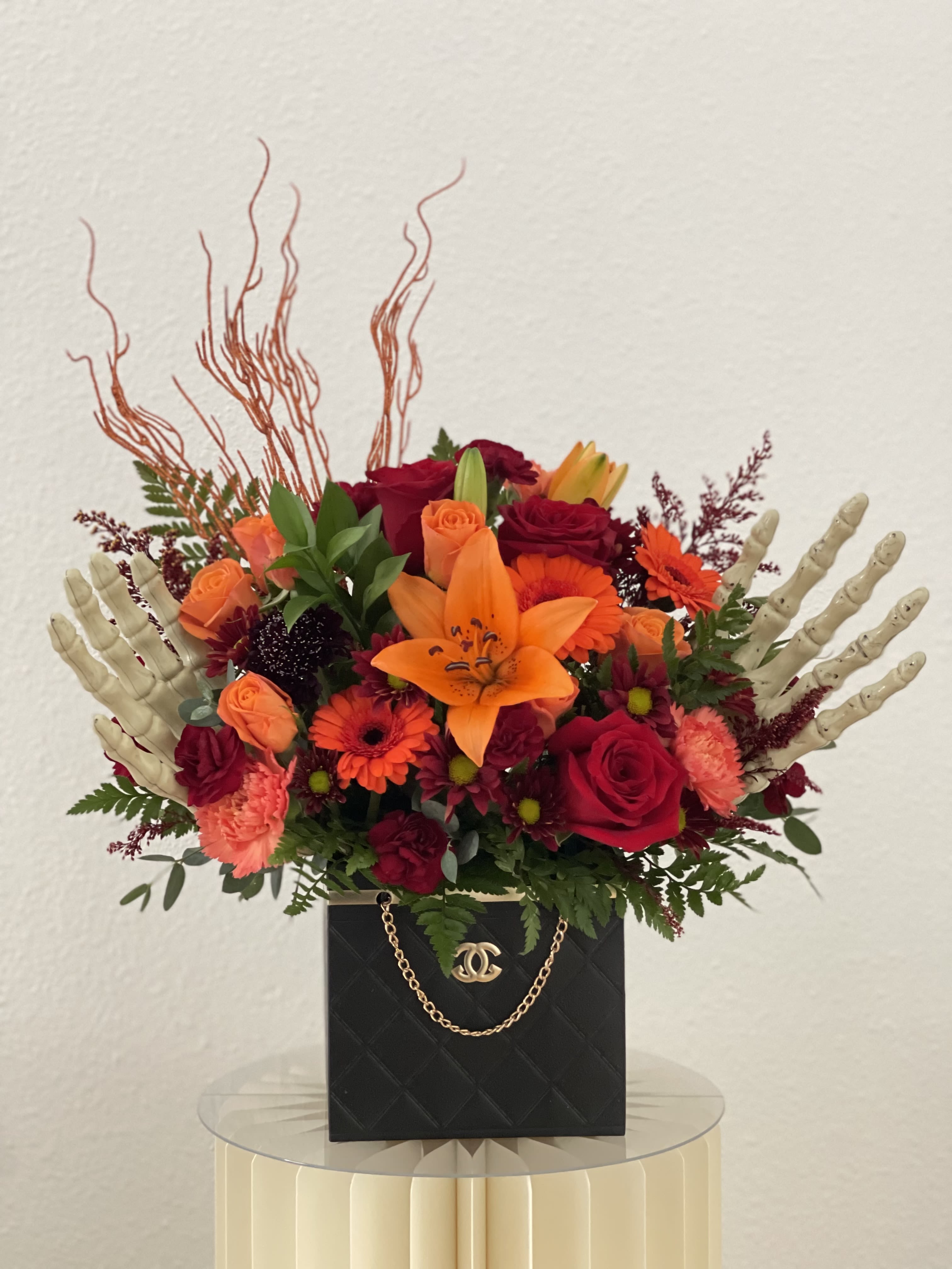 Bones &amp; Blooms - Introducing our Bones &amp; Blooms floral arrangement – a unique blend of red and orange roses, gerberas, carnations, chrysanthemums, and scabiosa, all nestled in a black box with skeleton hands peeking out. This quirky and playful display combines vibrant fall colors with a touch of spooky fun, making it a standout piece for Halloween or any time you want to add a whimsical twist to your decor. The perfect mix of cheerful blooms and whimsical details, our Bones &amp; Blooms arrangement is sure to bring a smile to anyone's face.