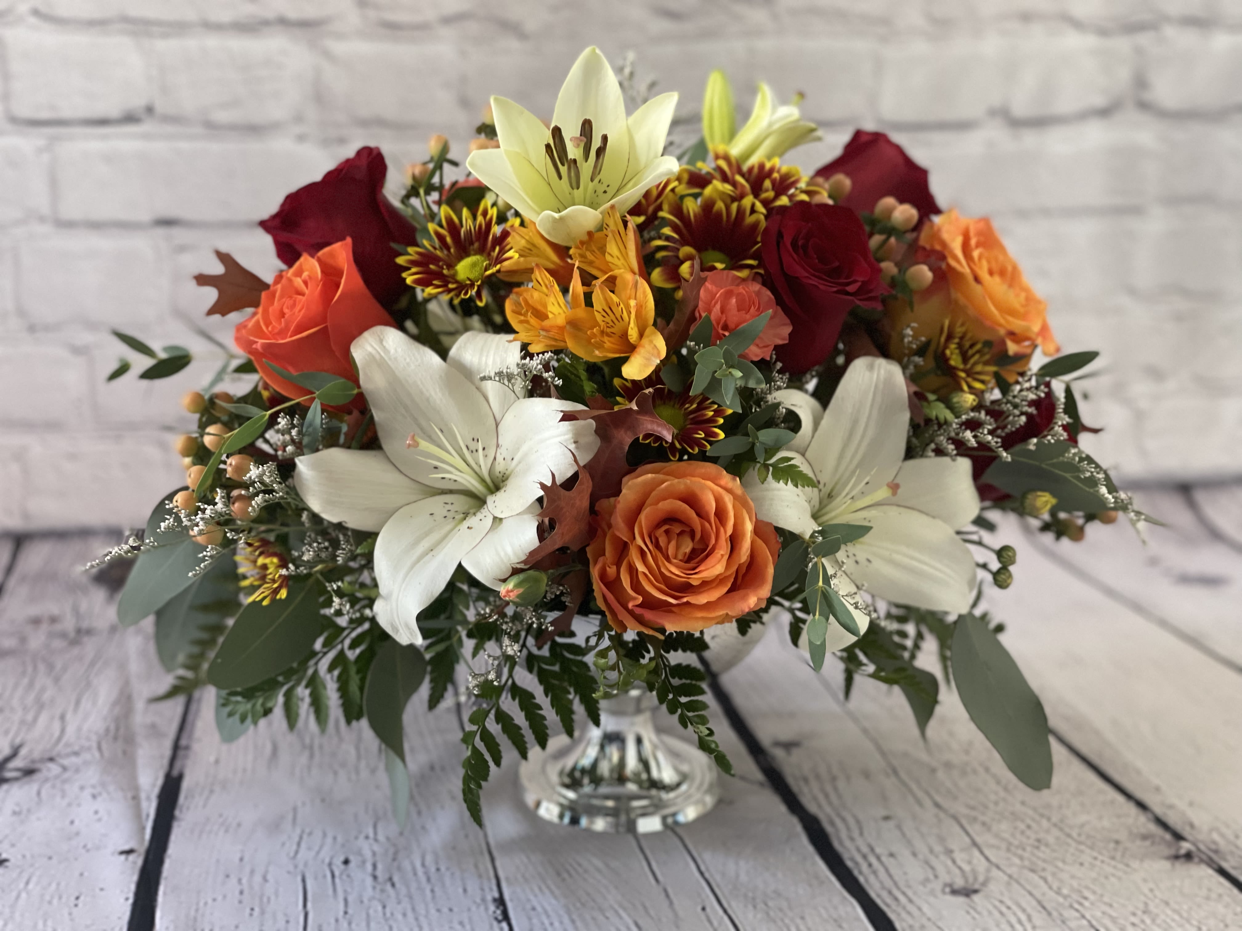 Gratitude Centerpiece - Celebrate the spirit of Thanksgiving with our exquisite Gratitude centerpiece, an opulent display of orange and red roses, lilies, and an array of autumnal flowers and foliage elegantly arranged in a gold or silver pedestal bowl. Radiating warmth and abundance, this stunning floral arrangement embodies the essence of gratitude and harvest, making it a captivating focal point for your Thanksgiving table. The rich colors and lush textures capture the spirit of the season. At the same time, the shimmering pedestal bowl adds a touch of elegance and sophistication, creating a truly unforgettable centerpiece for your festive gathering.