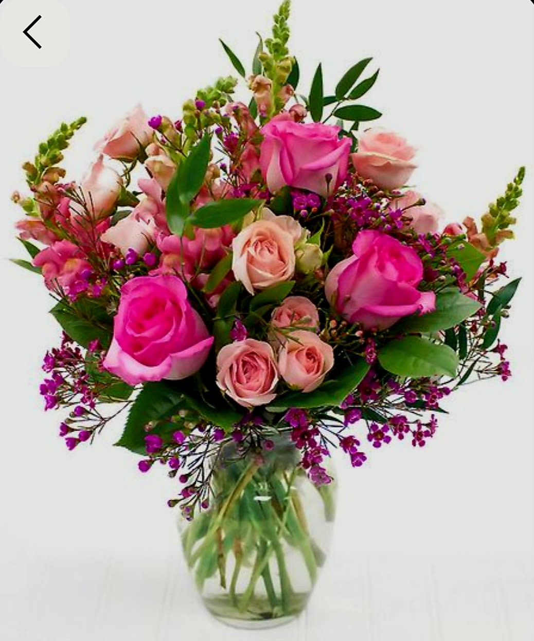 Friendship by Pretty in Petals  - This arrangement features a harmonious blend of pink and magenta roses, interspersed with clusters of small purple filler flowers and framed by lush green foliage. The roses range from soft blush tones to deep magenta, offering a gradient of color that adds depth and visual interest. The bouquet is densely packed and symmetrically styled, with each element carefully placed to create a balanced, outward-reaching form. Presented in a clear glass vase, the composition emphasizes freshness and elegance, making it suitable for celebratory settings or refined interior decor