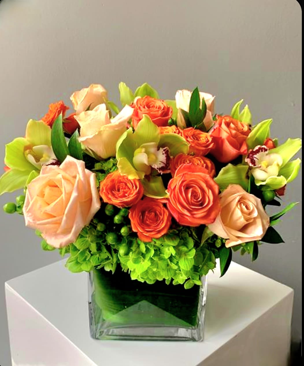 Peach Perfection by Pretty in Petals - This arrangement features peach and orange roses, green cymbidium orchids with maroon centers, green hydrangeas, and green hypericum berries. The flowers are compactly arranged in a clear square glass vase, accented with leafy greens for texture. The palette blends warm and cool tones, creating a balanced and vibrant composition.
