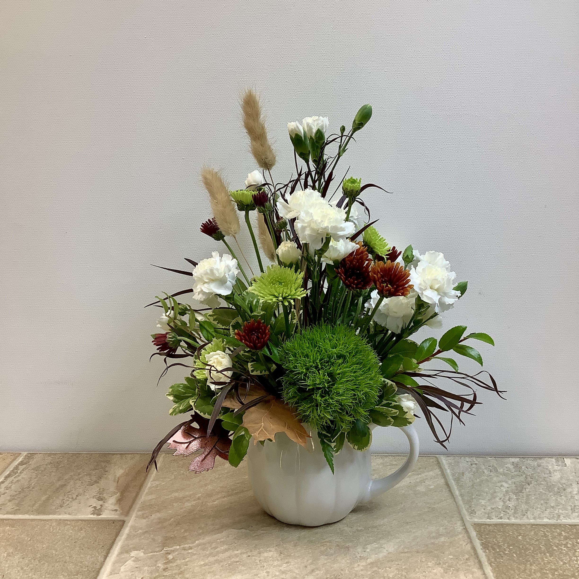Pumpkin Spice Pumpkin Spice - Bring the warmth of the season indoors with this charming autumn-inspired arrangement. Crisp white carnations, vibrant green mums, and rich burgundy accents are paired with soft bunny tail grasses and lush greenery. Designed in a cozy mug-shaped vase with a touch of autumn foliage, this piece captures the rustic charm of a fall meadow. Perfect for a seasonal centerpiece, a thoughtful gift, or simply to add a touch of autumn glow to your home. It measures 11” wide x 14” tall.