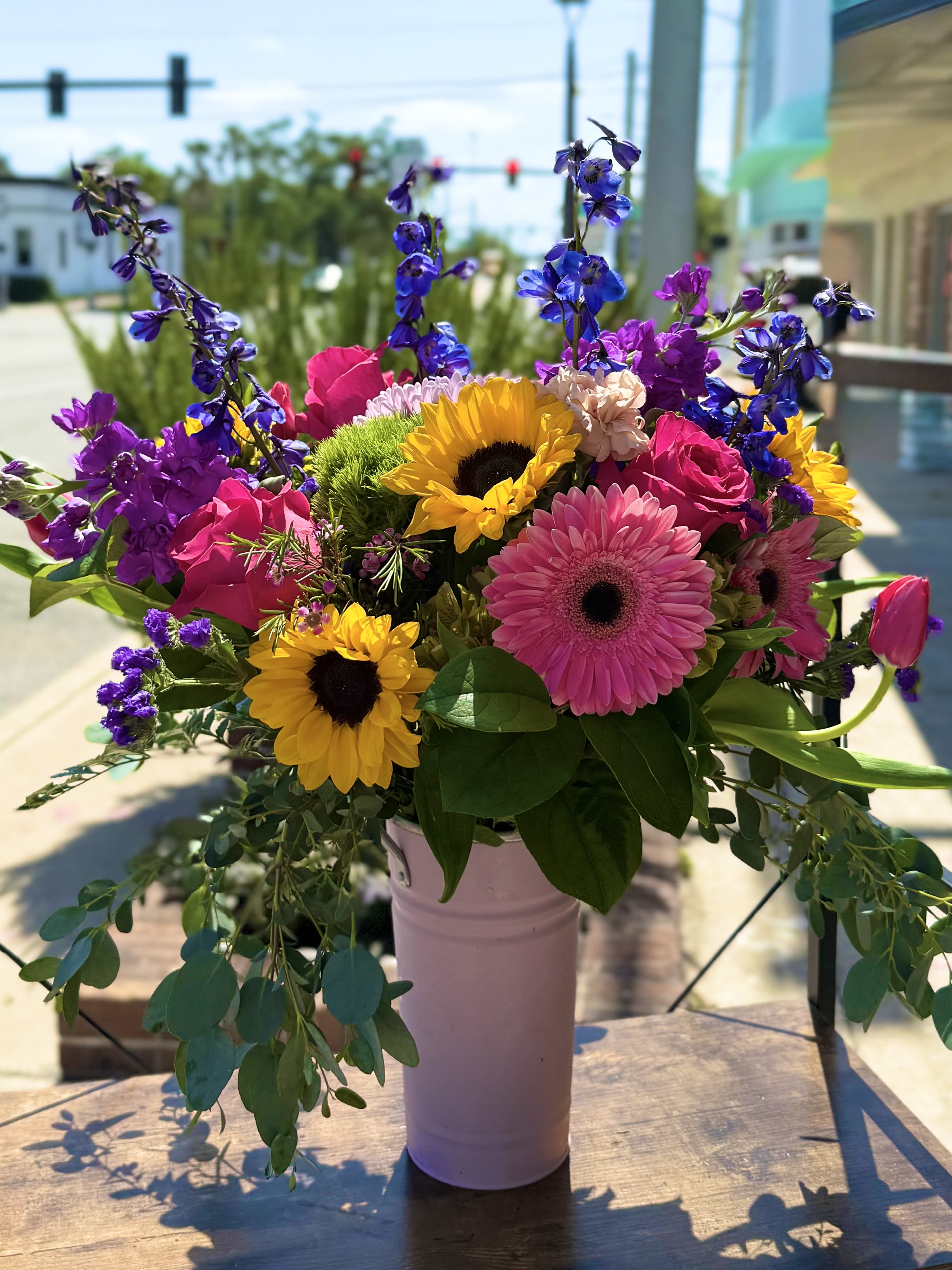 From Mom's Garden From Mom's Garden - Colorful blooms dripping from our keepsake baby pink metal vessel featuring Florida sunflowers, hot pink roses, rich blue delphinium, fragrant purple stock tulips and much more!