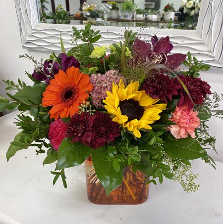 Autumn Glow Autumn Glow - This cheerful autumn arrangement radiates warmth and charm. A golden sunflower and bright orange gerbera daisy take center stage, surrounded by rich burgundy mums, deep-toned orchids, and soft peach carnations. Accents of green and textural foliage add balance and freshness, while the autumn-hued vase ties the whole design together. Perfect for celebrating the season, this bouquet brings a vibrant glow to any table or occasion.