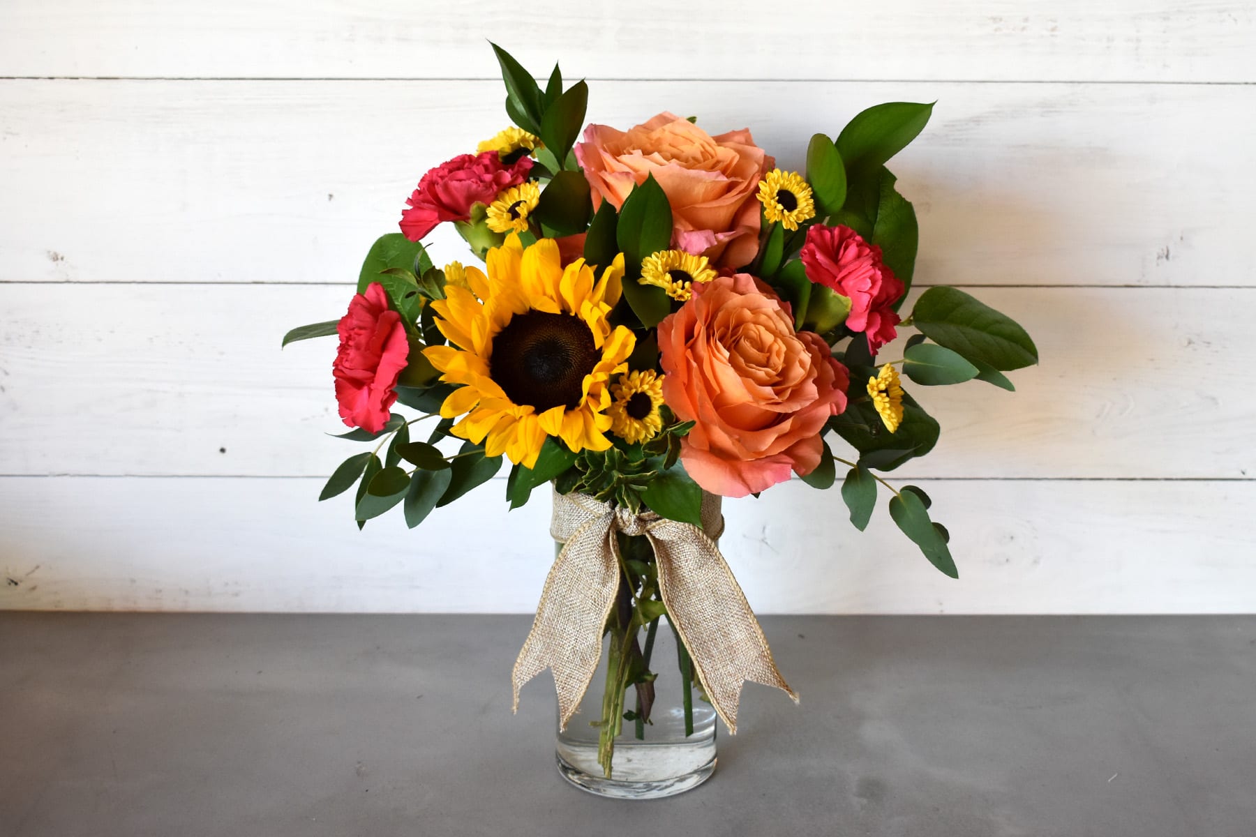 Stacey  -  This arrangement features a mix of orange roses, carnations, sunflowers, greens and filler in a  glass vase.