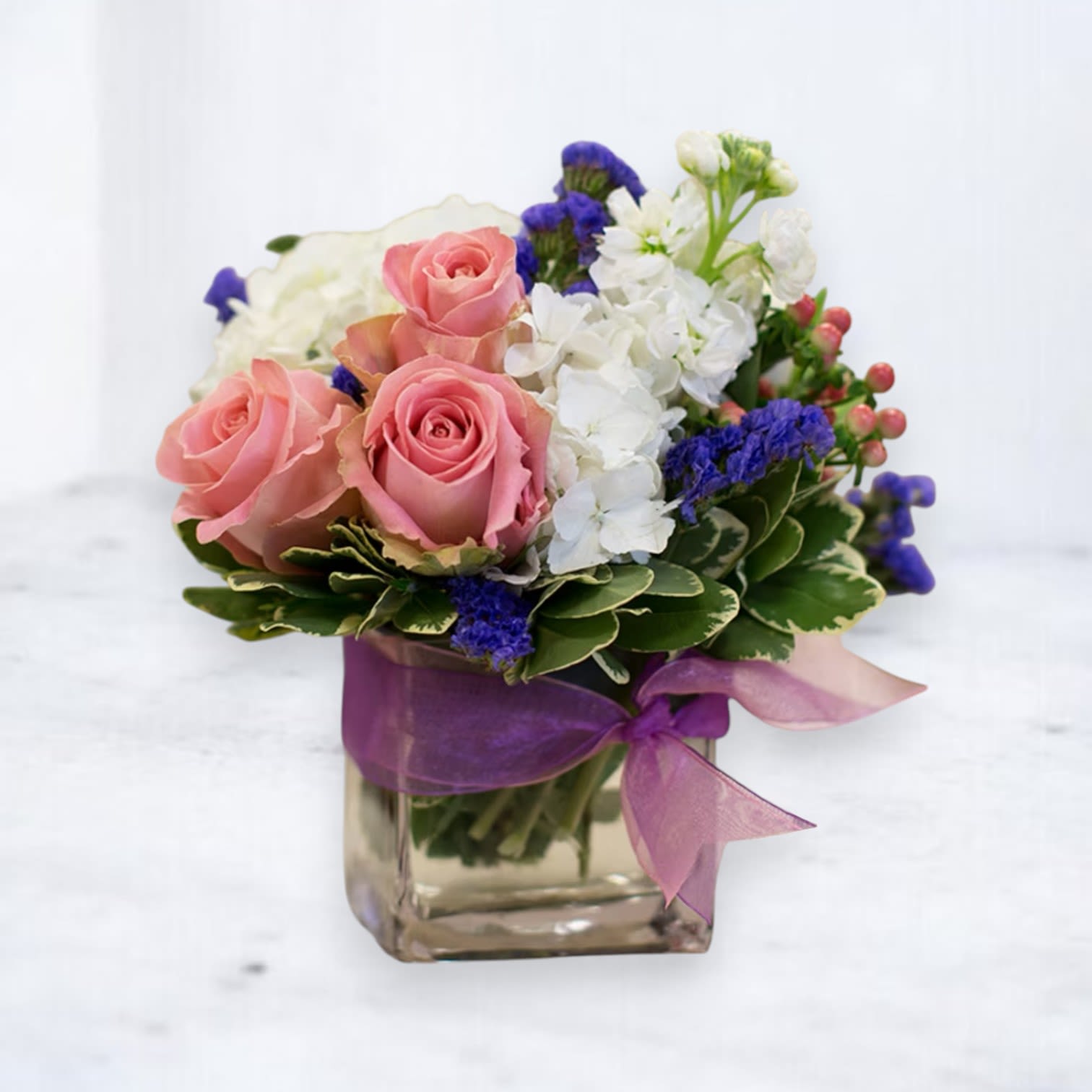 Lavender Whisper Charm - Delight in the enchanting beauty of the Lavender Whisper Charm, a compact yet captivating floral arrangement that exudes timeless elegance. Featuring soft pink roses, pristine white hydrangeas, vibrant purple statice, and delicate hypericum berries, this bouquet is accented with lush greenery and tied with a luxurious purple ribbon for a finishing touch.  Perfect for birthdays, thank-yous, or simply brightening someone’s day, this arrangement is a charming gift that speaks volumes. Available for same-day delivery in Houston and surrounding areas, the Lavender Whisper Charm is a thoughtful and stylish choice for any occasion.