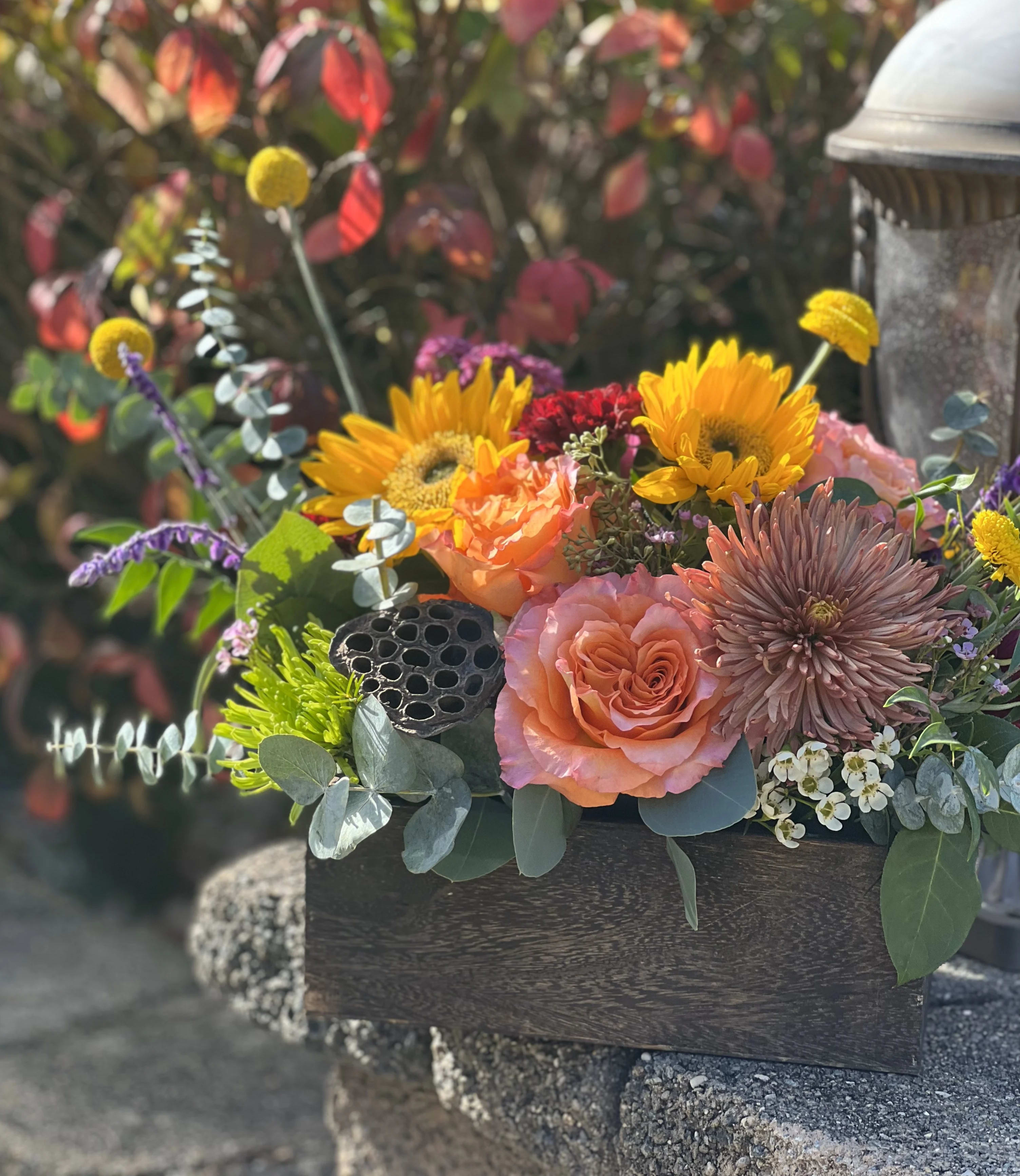 Falling Leaves - A vibrant rustic arrangement to usher in the changing of the leaves and crisp cool weather. This design is arranged in a handmade wooden box with an assortment of fall blooms- sunflowers, mums, roses, eucalyptus and a variety of seasonal textures.