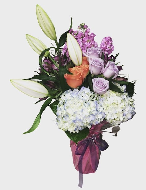 Pink and Yellow Arrangement - Orange and Lavender roses, Lavanda Stock, White Lily, Bupleurum, Limonium, Purple and Orange Alstroemerias, Lemon Leaf, Bupleurum, Greens. Clear Vase 10” H