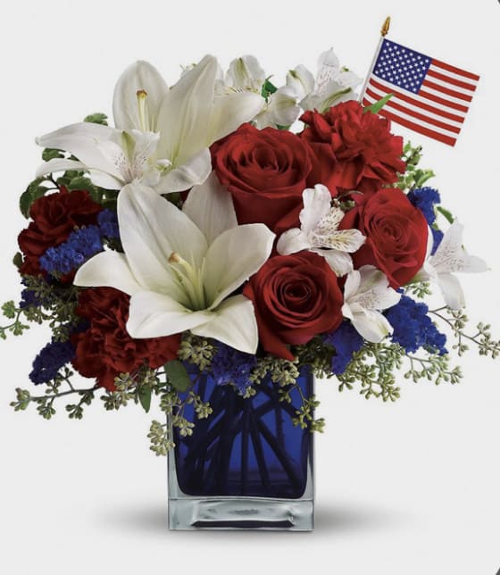 Patriot Arrangement Flowers - Red roses and carnations, white lilies and Blue Chrysanthemums, fillers and greens. Blue Square vase 4”x 4” and Small USA Flag.
