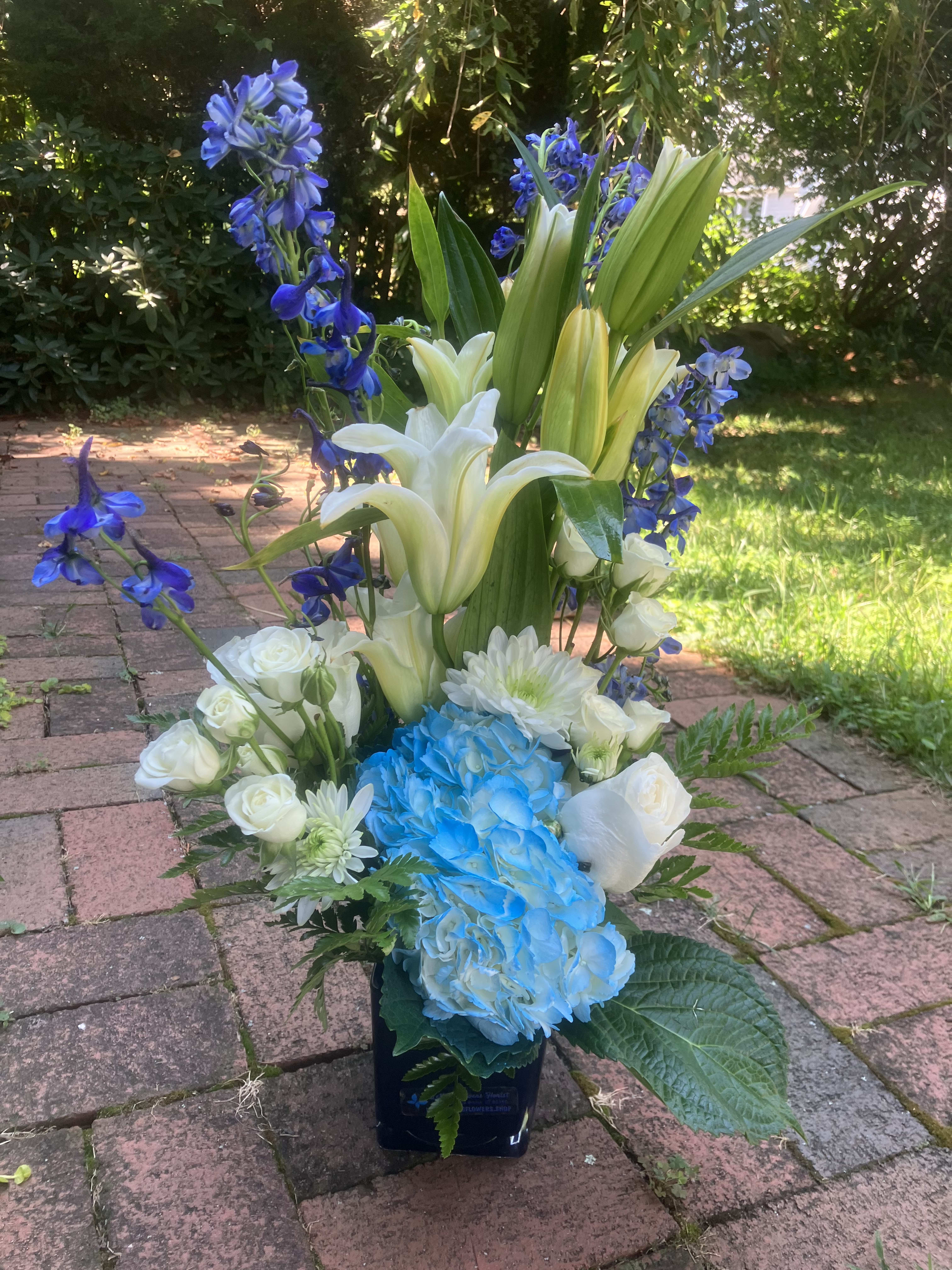 Sympathy Blue and White  - White Oriental Lily,White Roses, blue Hydrangea, Chysanthemum Spray green, Blue Delphiniums, and seasonal greens in a cute Blue square vase.