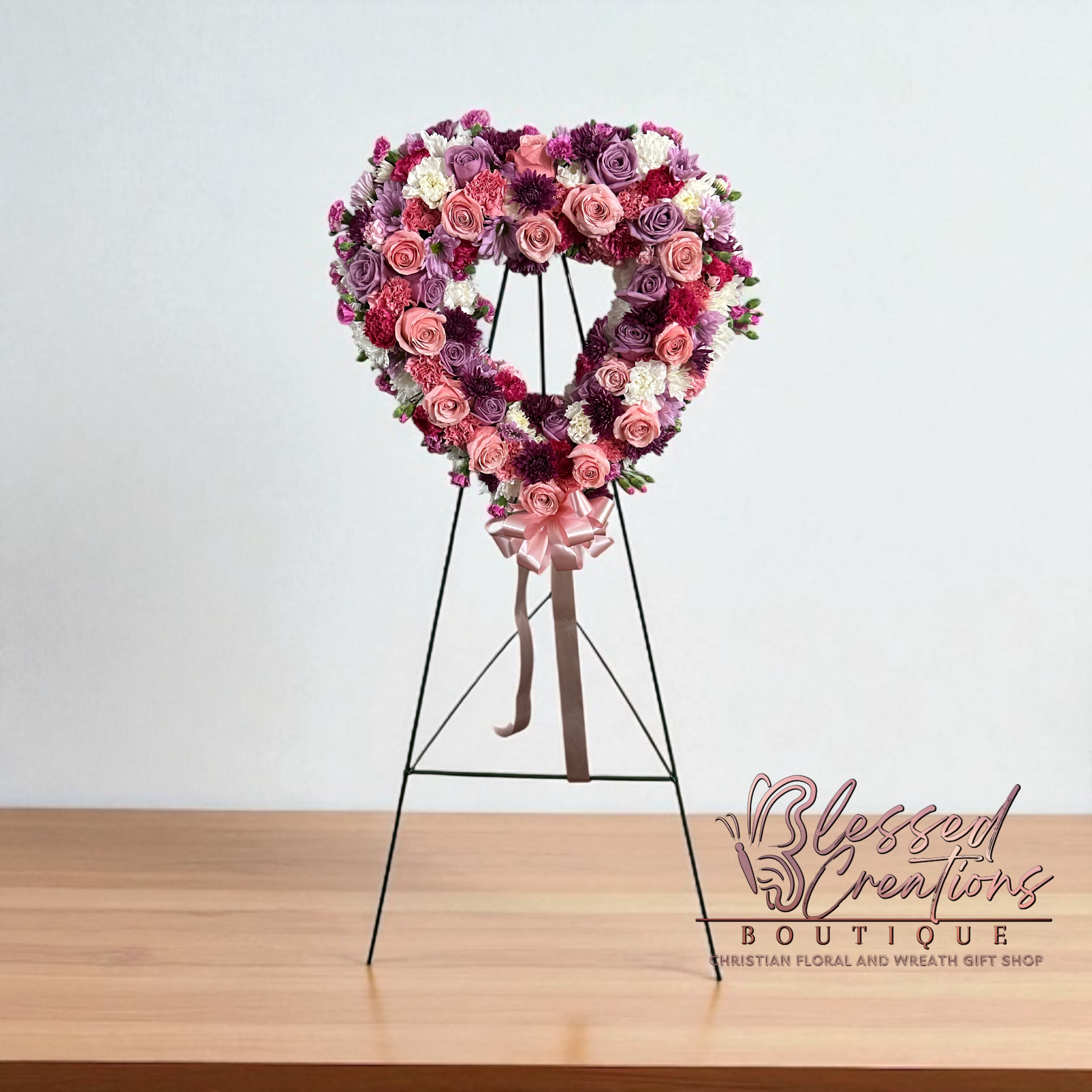 Sacred Love Tribute  - A beautiful tribute of love and remembrance. This heart-shaped wreath is designed with a graceful blend of lavender roses, soft pink roses, deep purple mums, carnations, white cushion mums, and accent greenery, finished with a delicate pink satin bow and streamers. A touching expression of sympathy and devotion, honoring cherished memories that will never fade.